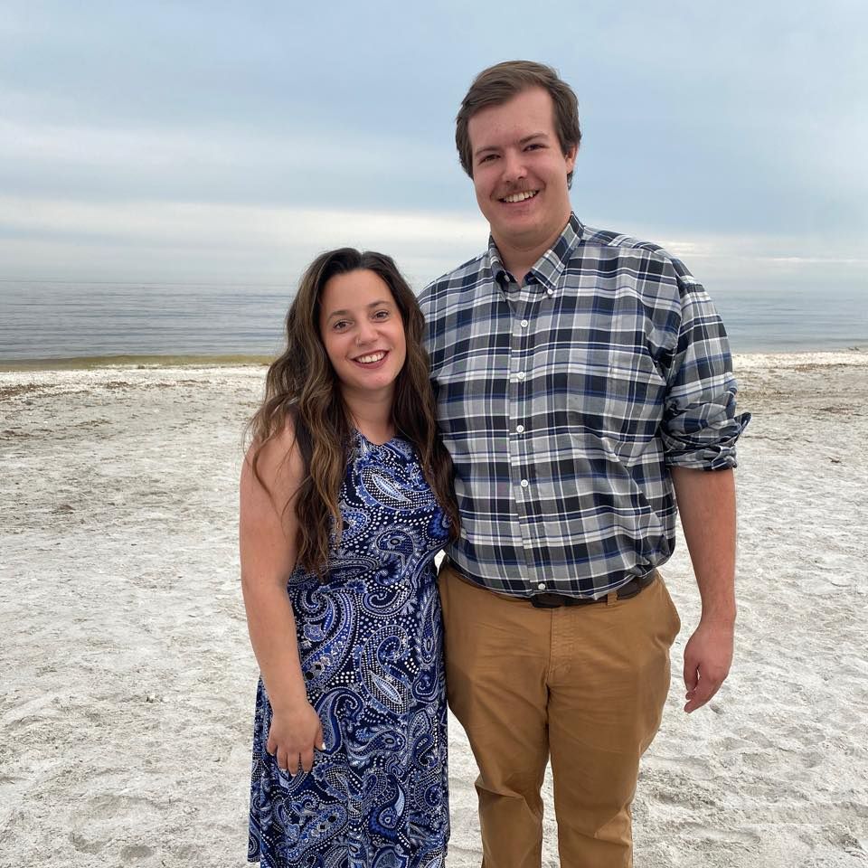 Couple in casual attire smiles while standing together on white sandy beach.