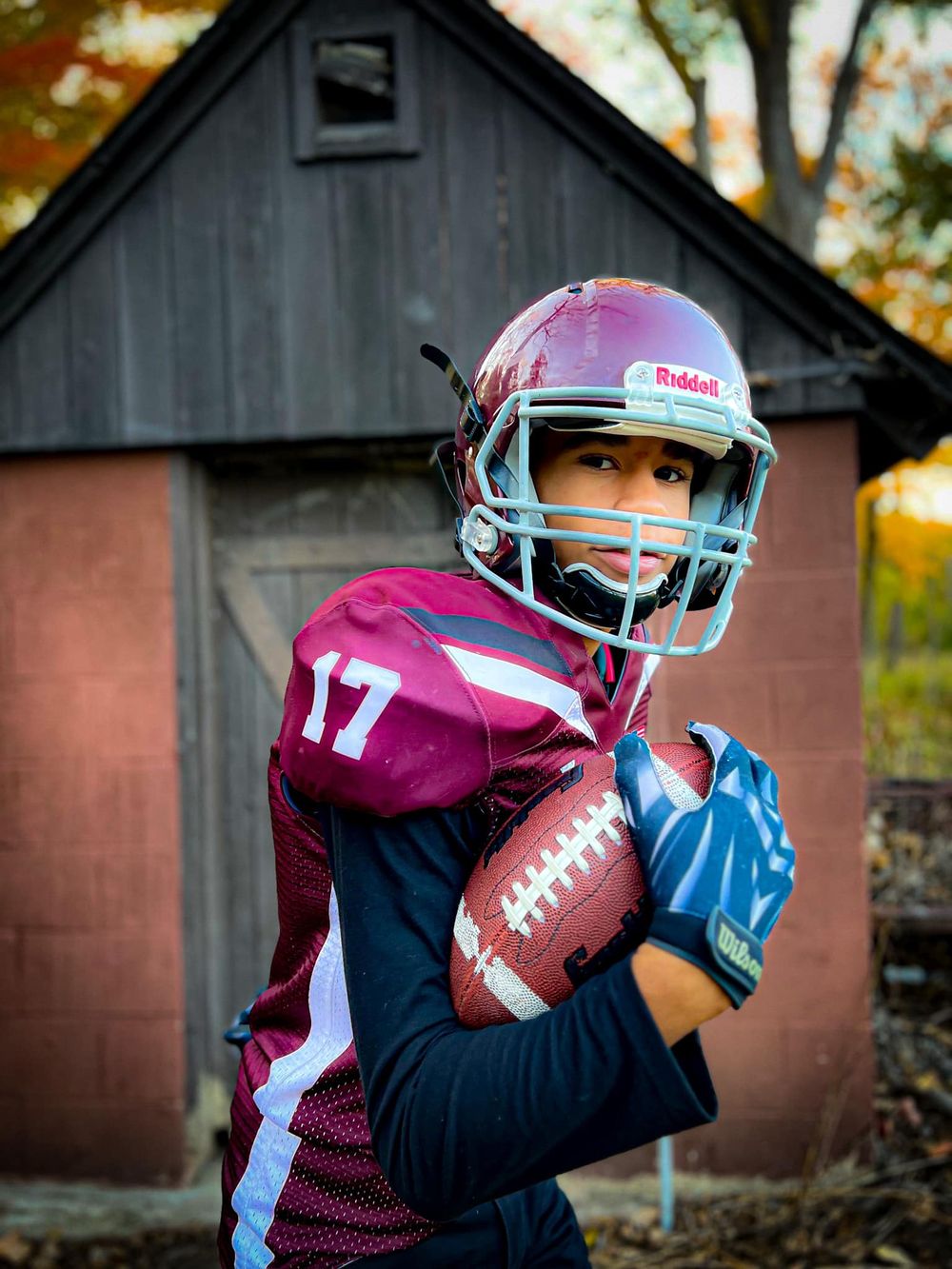A young football player in a maroon uniform poses with a ball in front of a rustic barn.