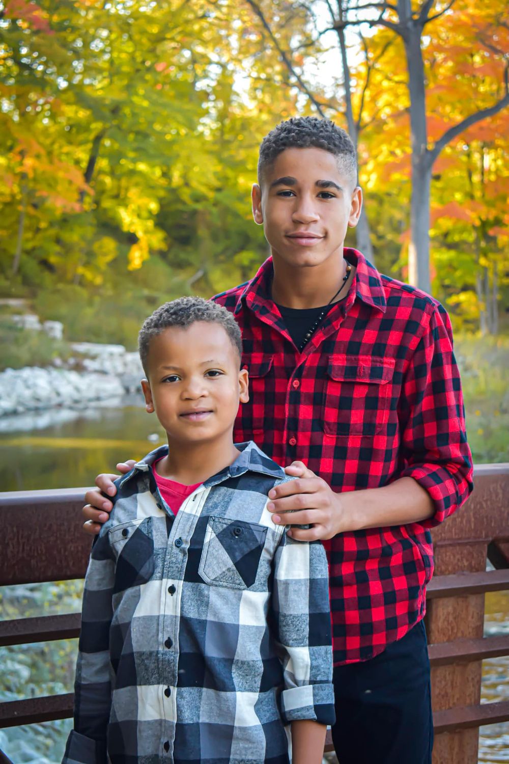 Two people in matching buffalo plaid shirts pose on a bridge with autumn colors in the background.