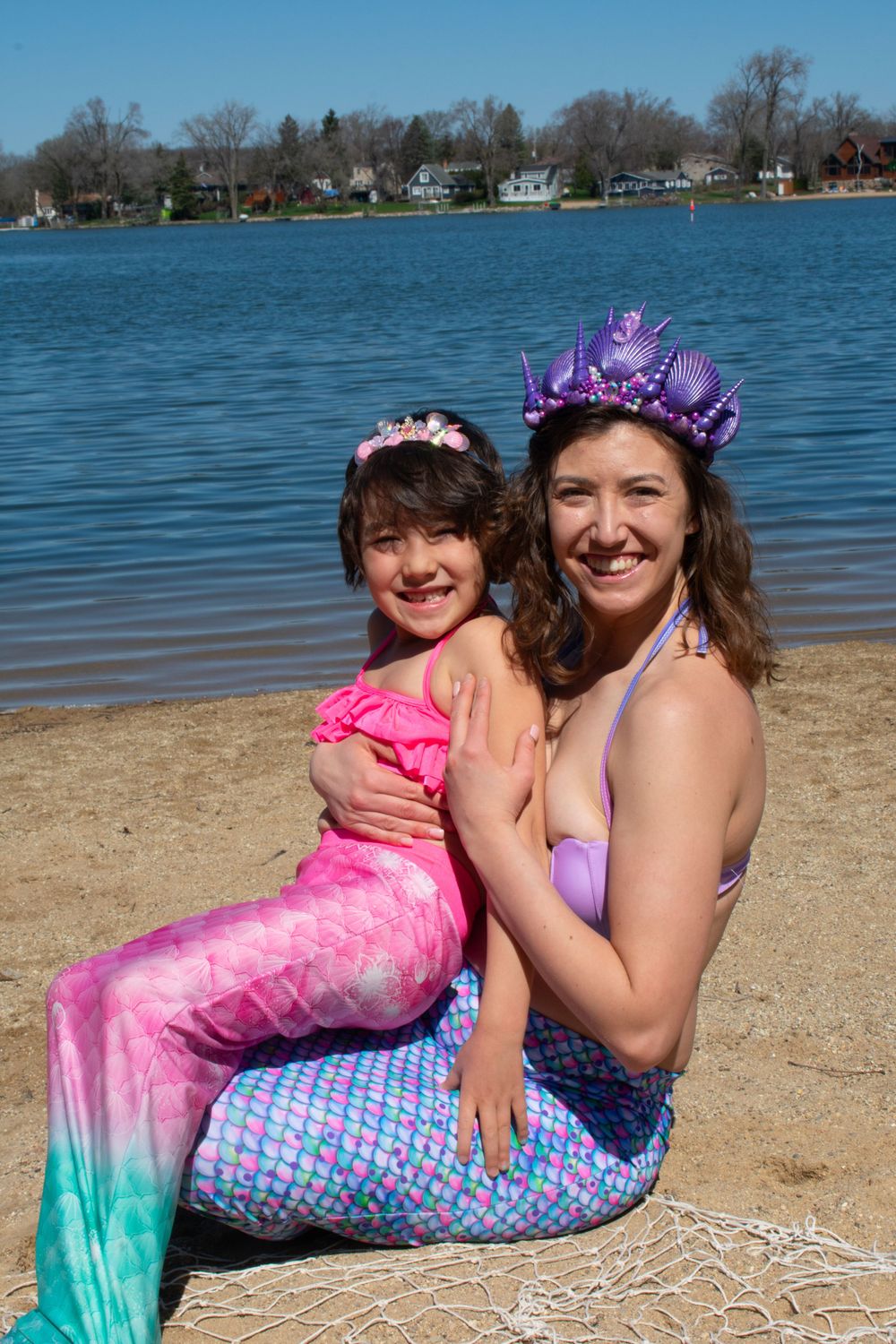 Two people in mermaid costumes pose together on beach beside lake.