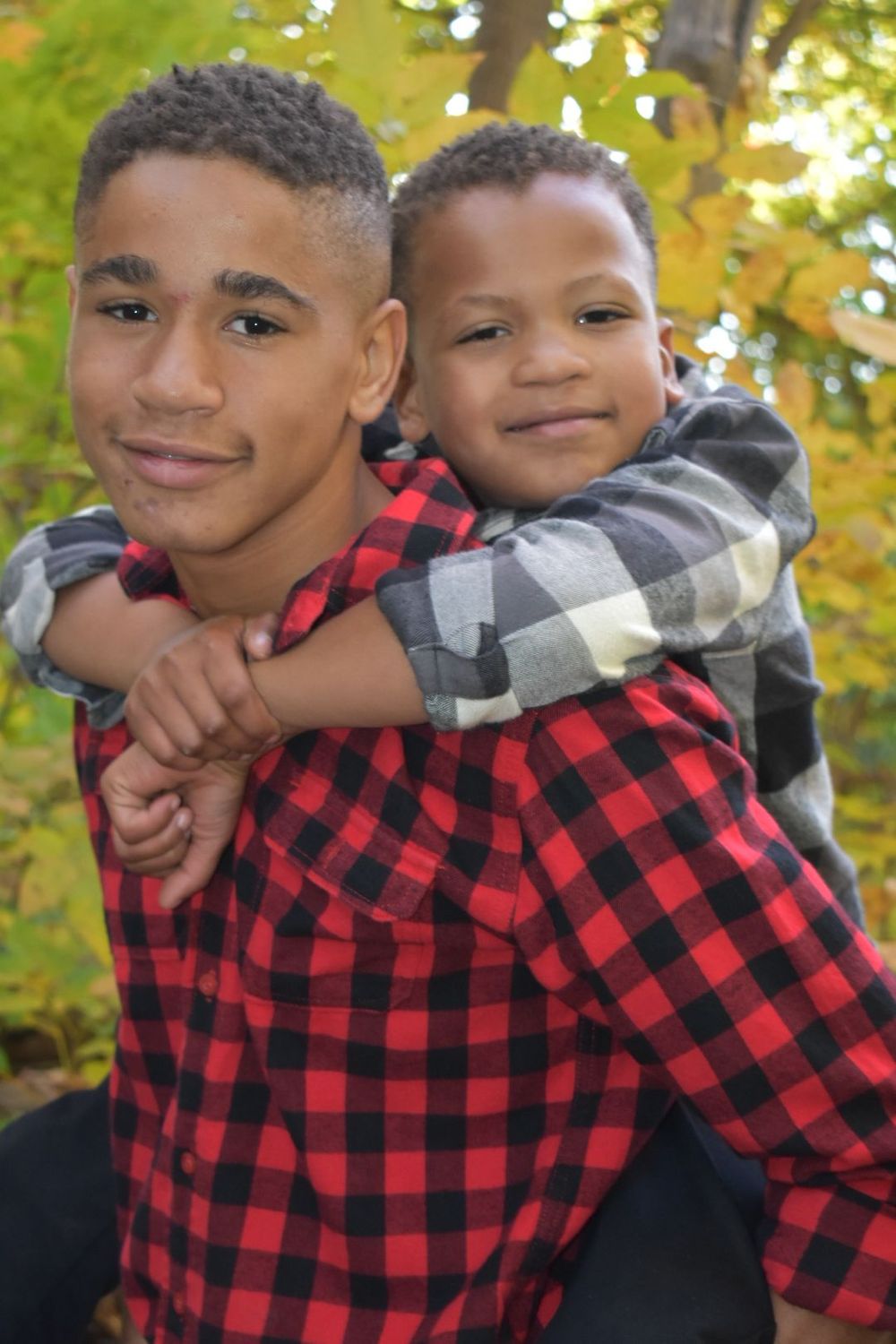 Two brothers in matching plaid shirts share playful piggyback ride in autumn setting.