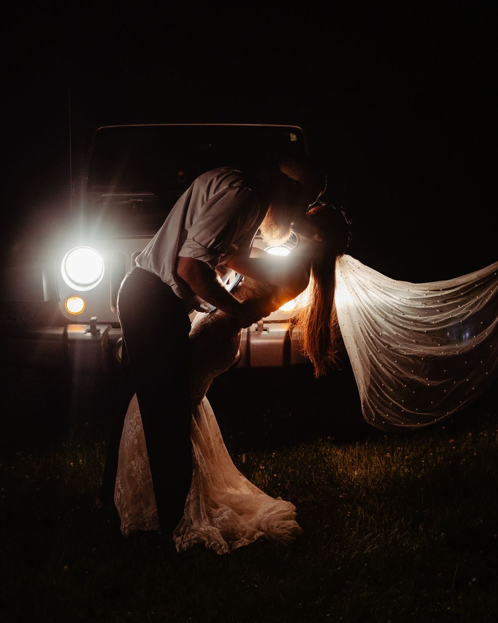 Romantic nighttime wedding portrait illuminated by vintage car headlights in a dramatic silhouette.