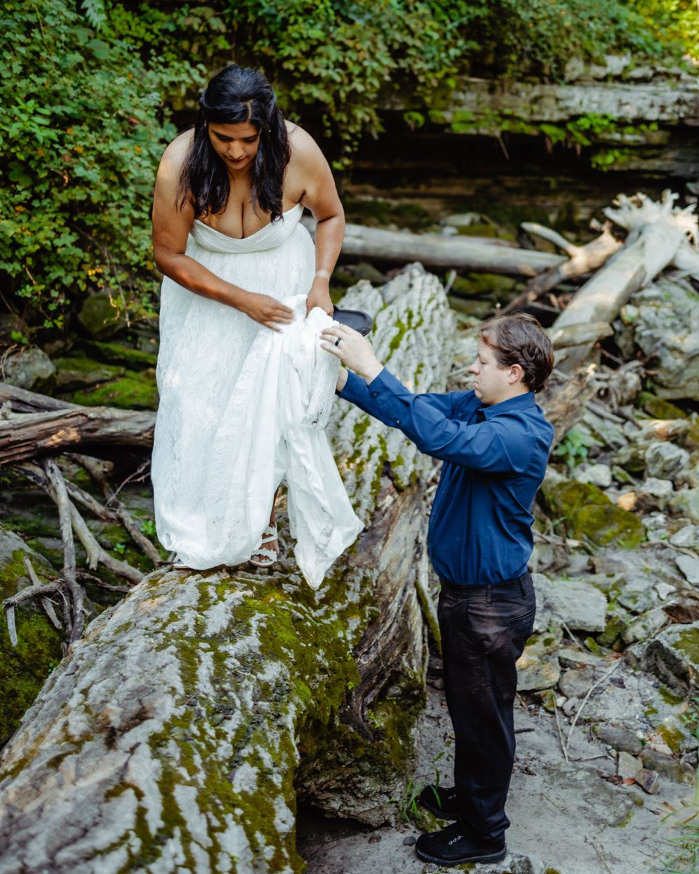 A bride in a white dress stands on rocks by a stream while assisted by someone in a blue jacket.