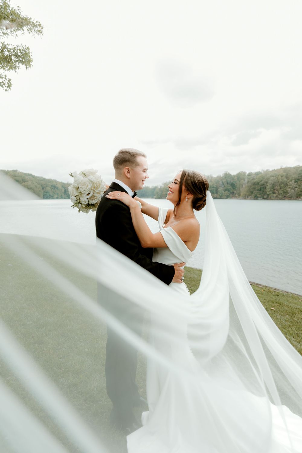 Regal husband and wife embracing lake side after their first look. Long Veil is pulled across the frame