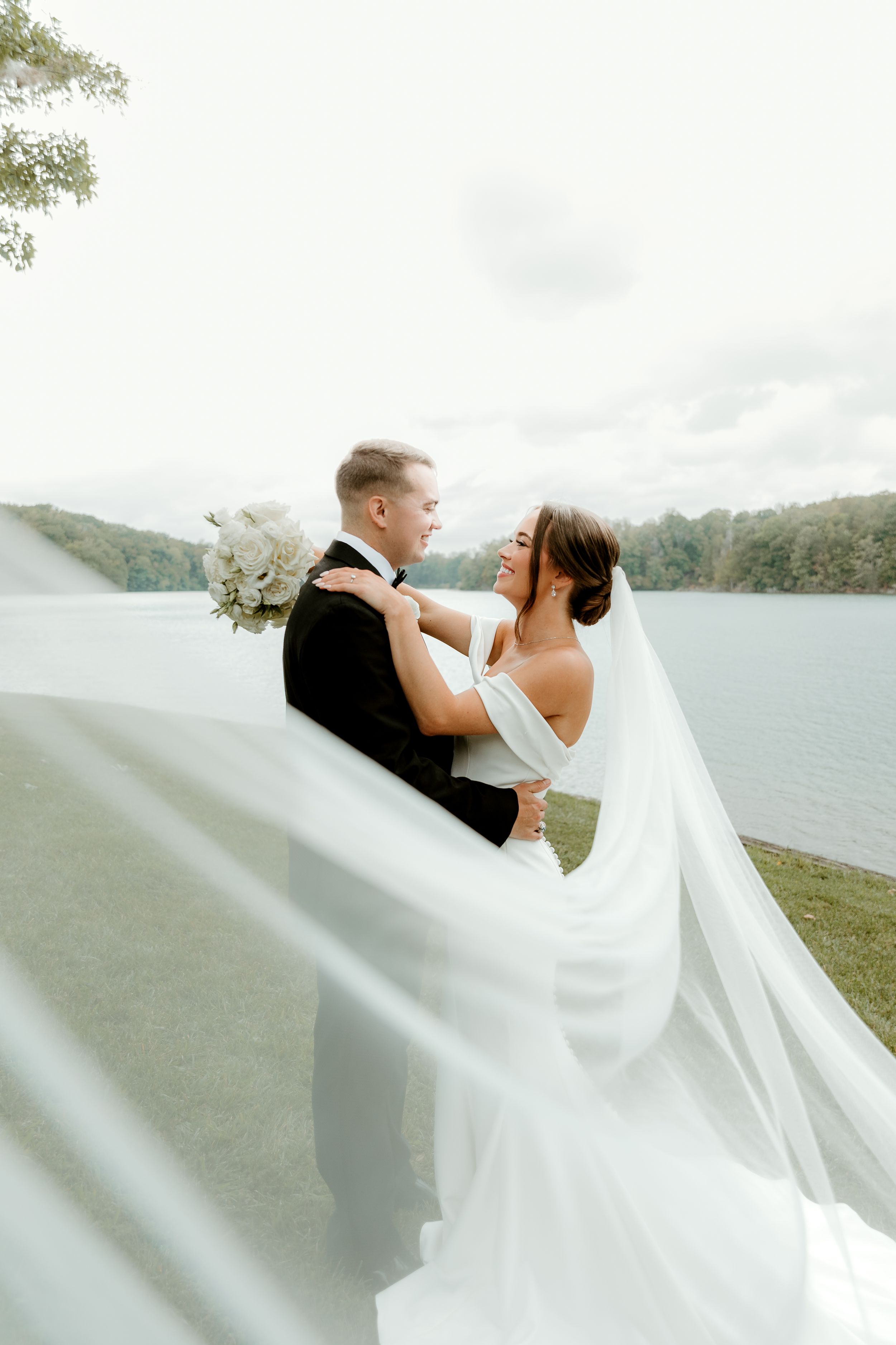 Regal husband and wife embracing lake side after their first look. Long Veil is pulled across the frame