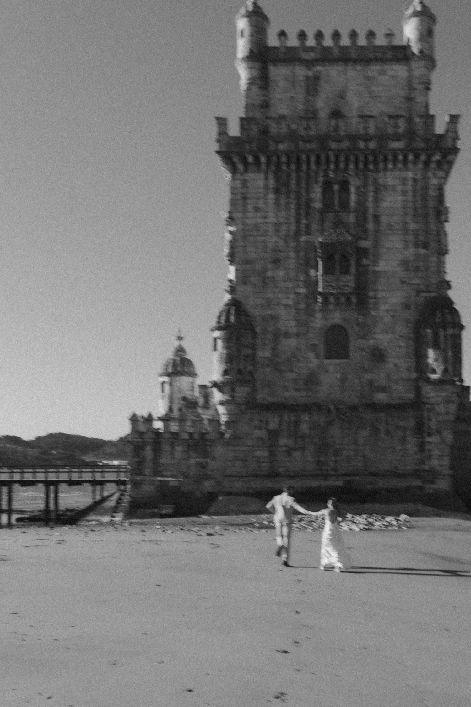 Black and white photo of Belém Tower in Lisbon with two figures walking on the beach in front of the historic fortress.