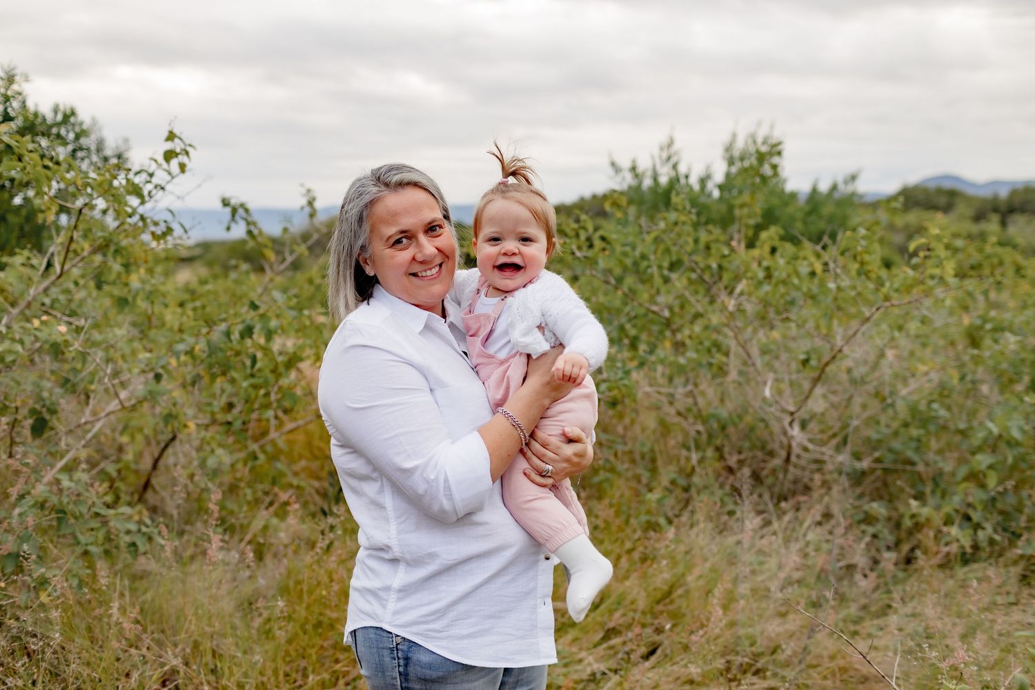 Parent holding smiling baby outdoors in field with mountains visible in background.