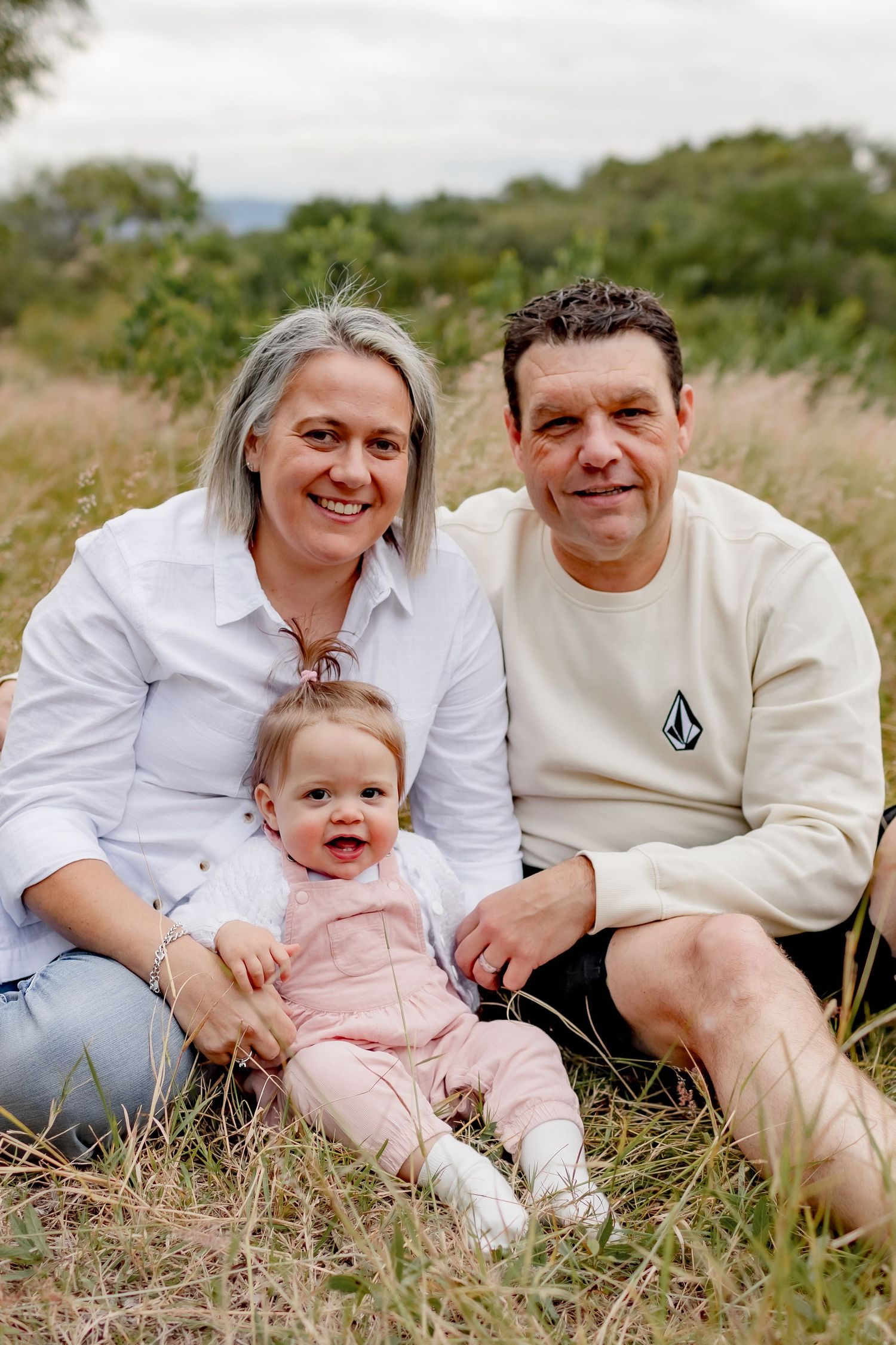 Casual family portrait with baby sitting together in tall grass on cloudy day.