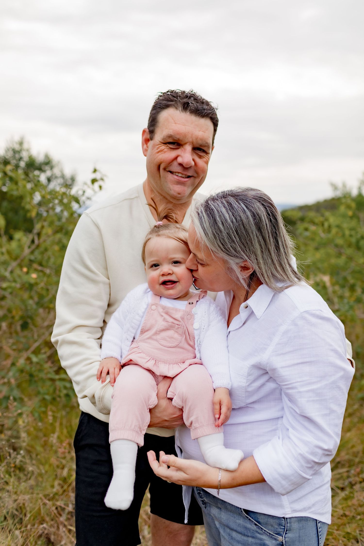 Family portrait outdoors in nature shows loving moment with baby and parents on cloudy day.