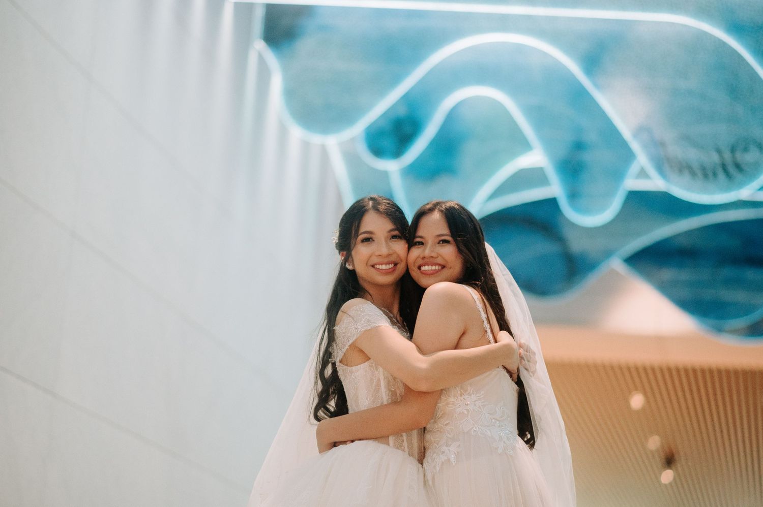 Two brides in white wedding dresses share a joyful embrace under modern blue wave ceiling decor.