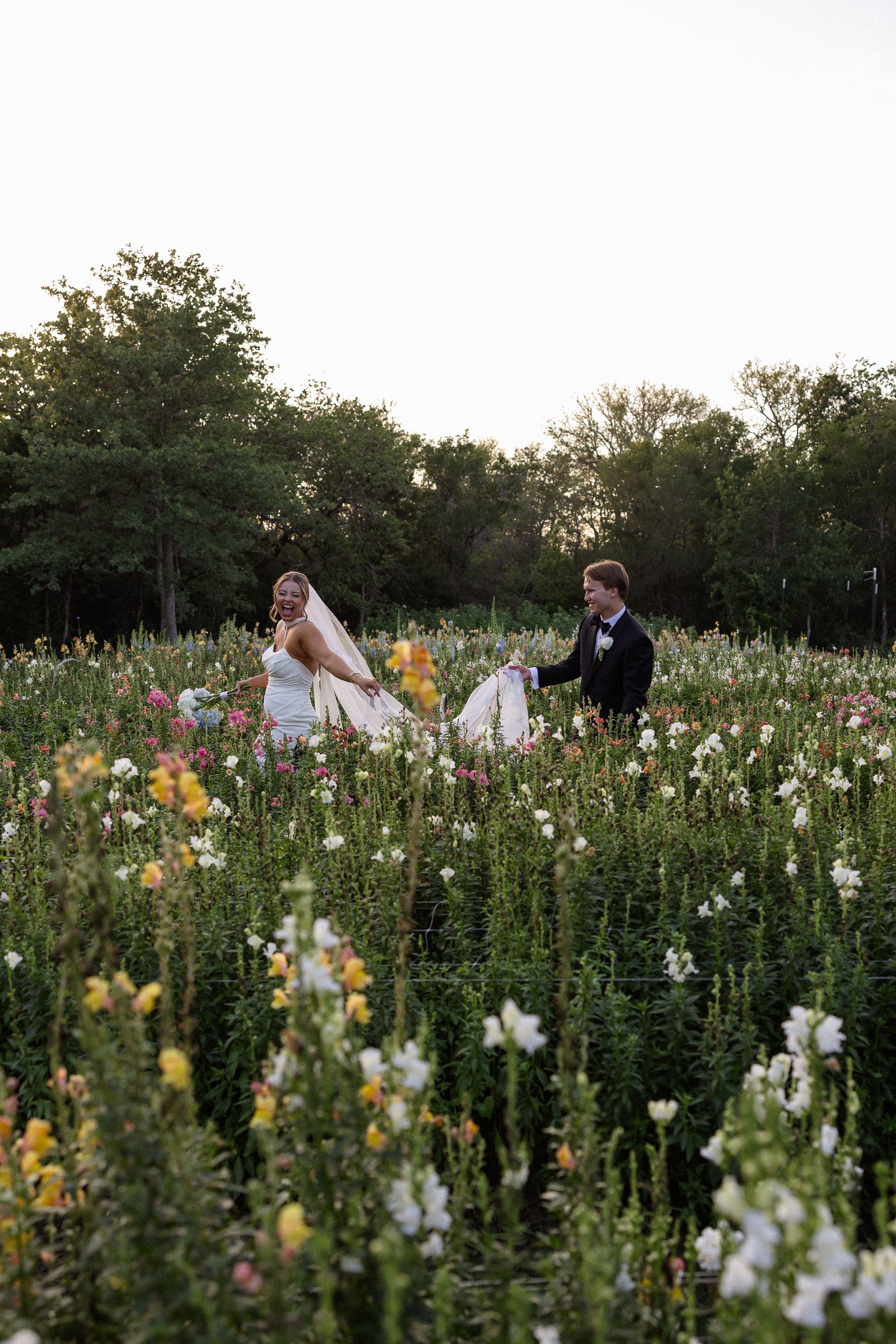 Bride and groom walking through a vibrant wildflower field at dusk, surrounded by colorful blooms and lush green trees.