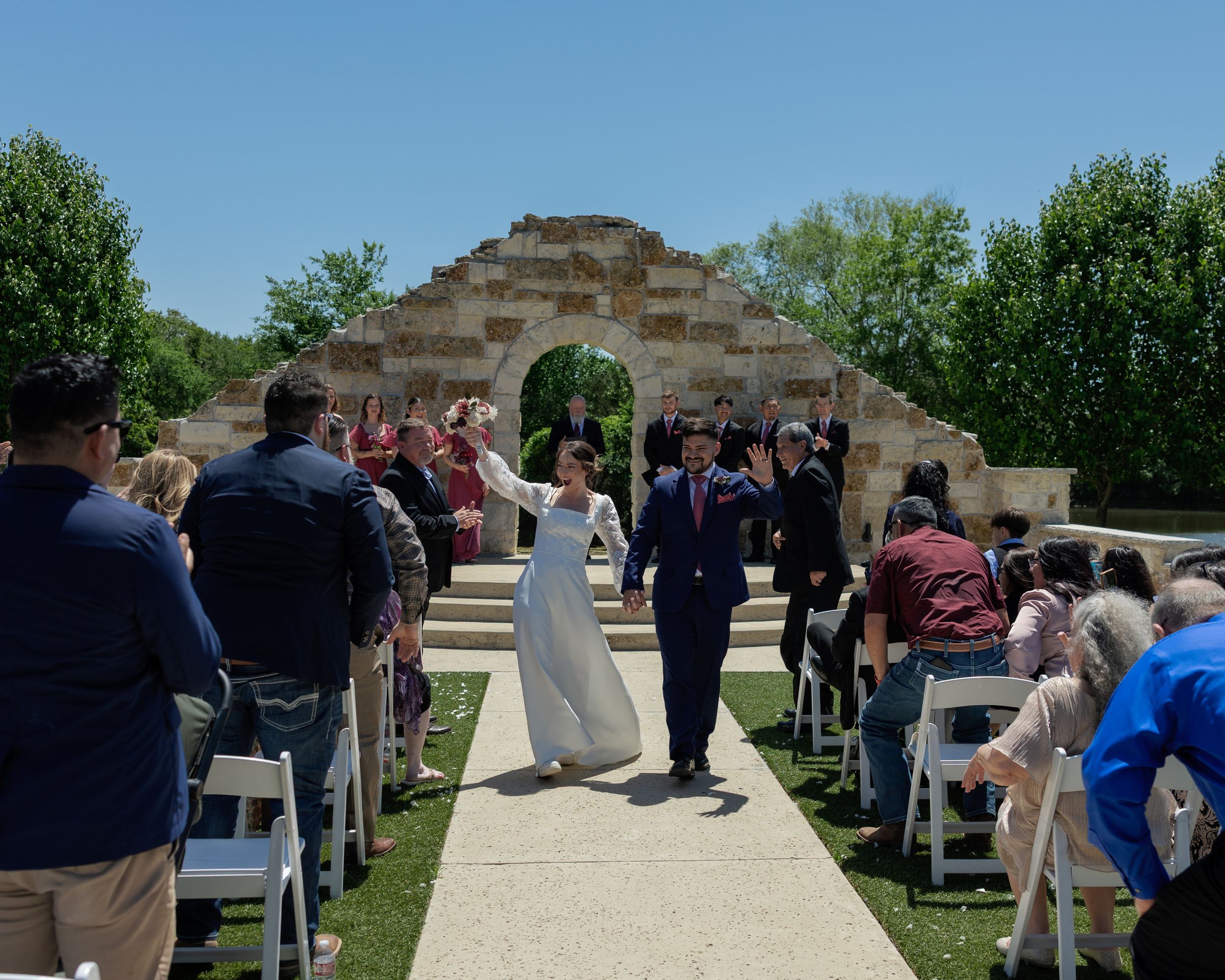 Outdoor wedding ceremony at stone archway with guests seated on white chairs along a paved pathway on sunny day.