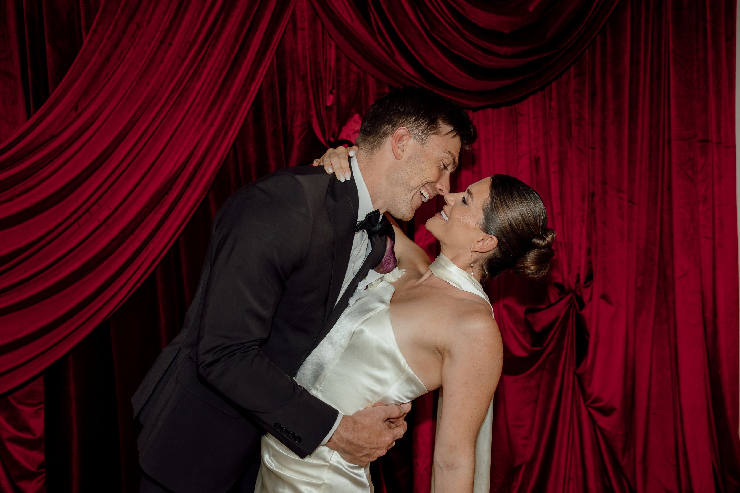Couple sharing a romantic kiss in wedding attire against a dramatic red velvet curtain backdrop.