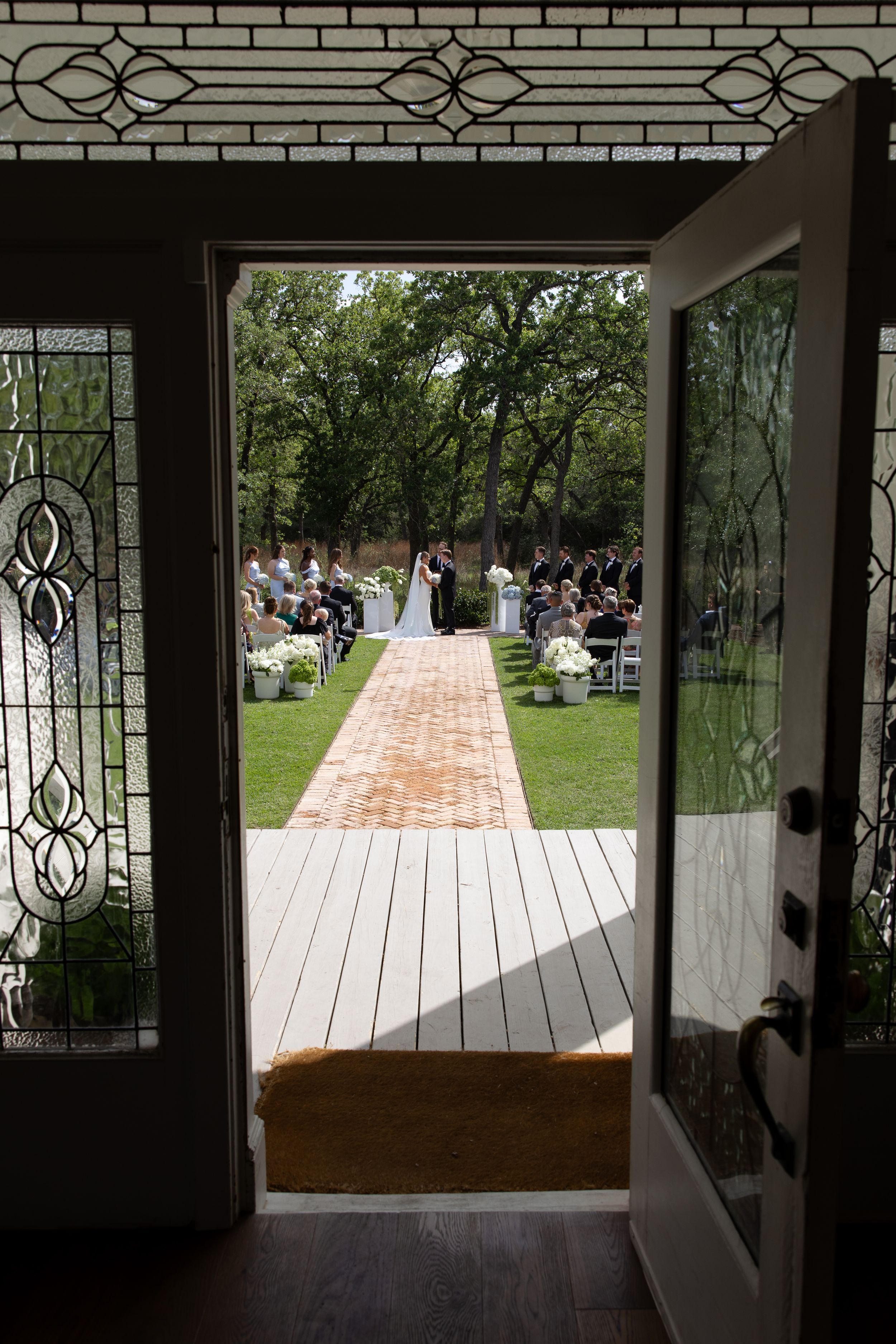 View through open doors of an outdoor wedding ceremony with couple at altar, lined aisle, and guests seated among lush green trees.