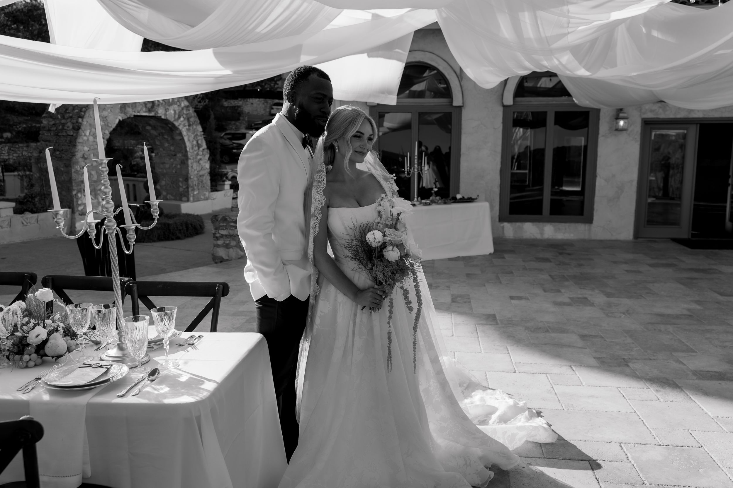 Black and white photo of a romantic outdoor wedding reception with elegant table settings and draped white fabric overhead.