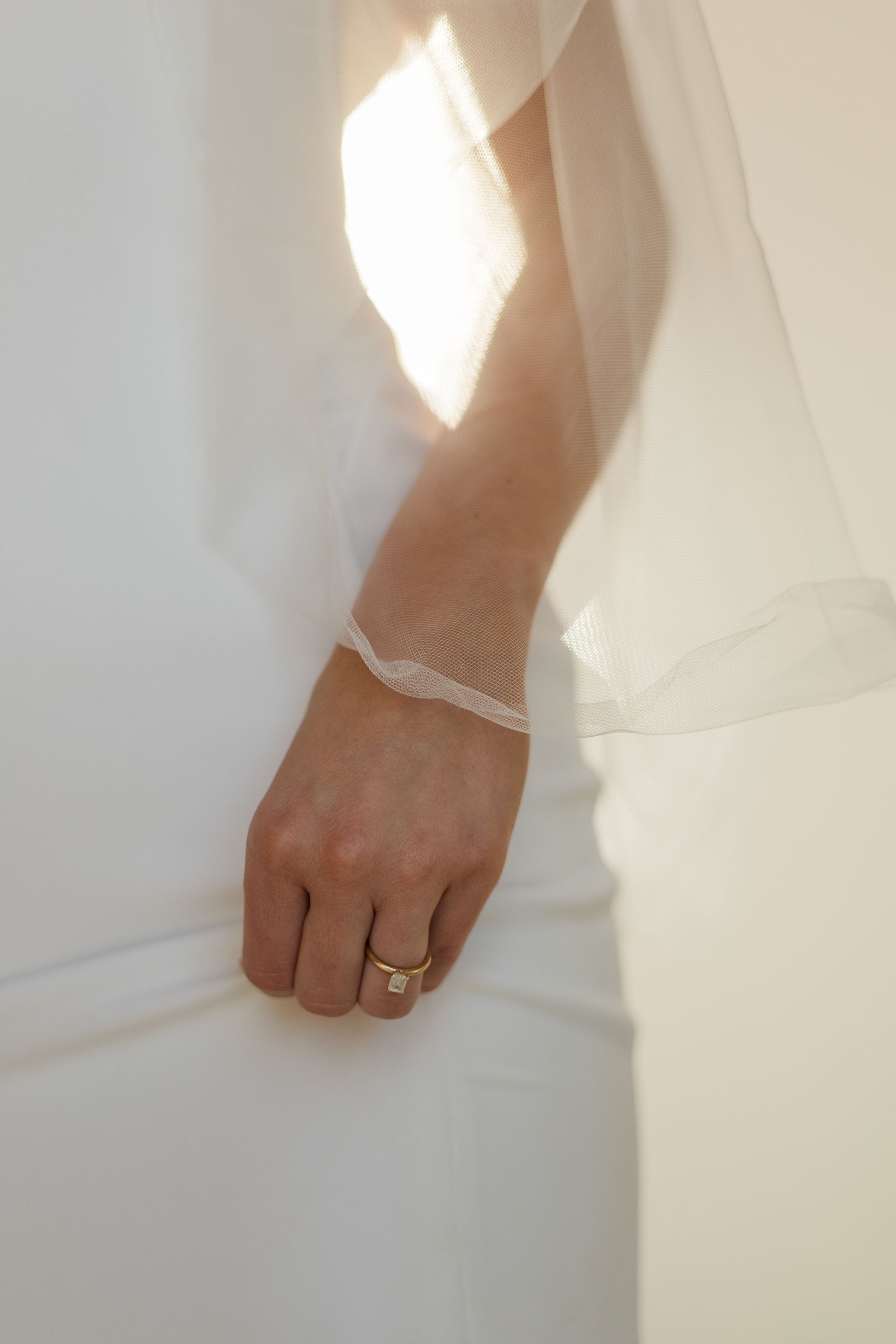 Artistic shot of wedding ring on hand against sunlit white dress.