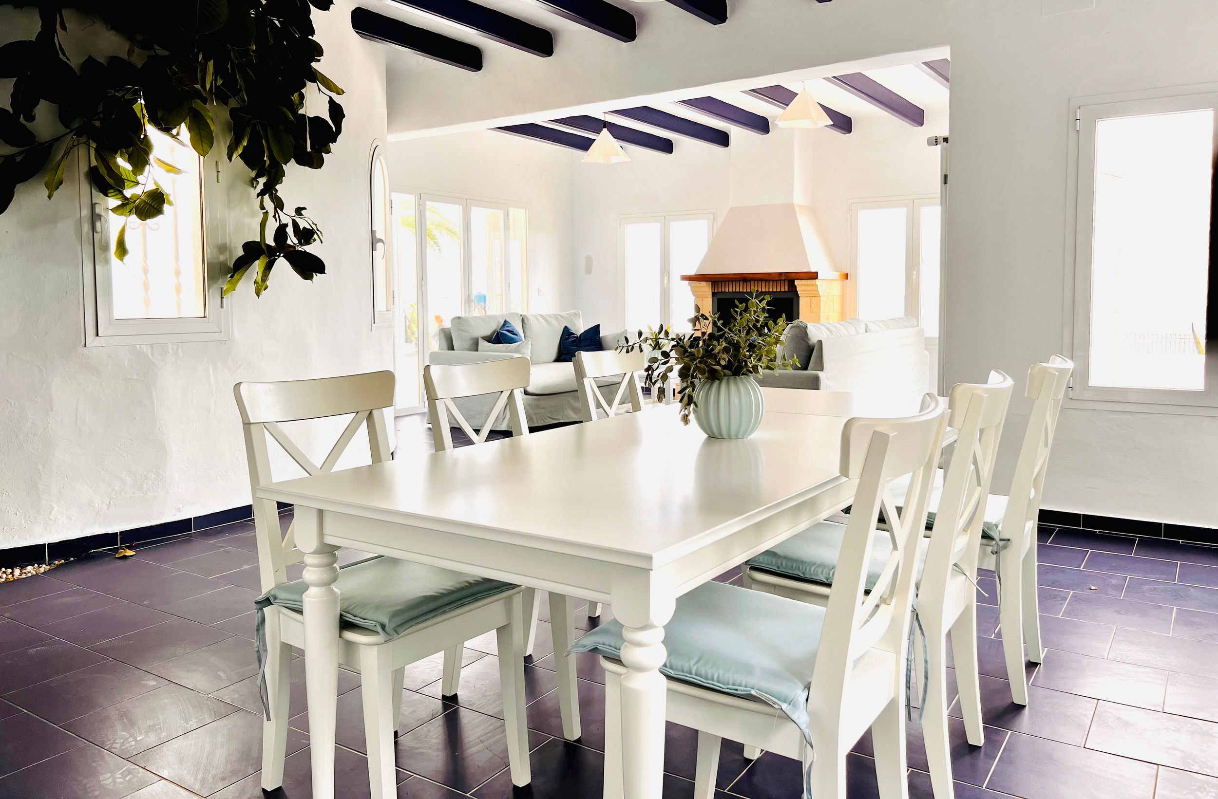 Elegant white dining room with farmhouse table, cross-back chairs, and dark tile floors in a bright sunlit space.