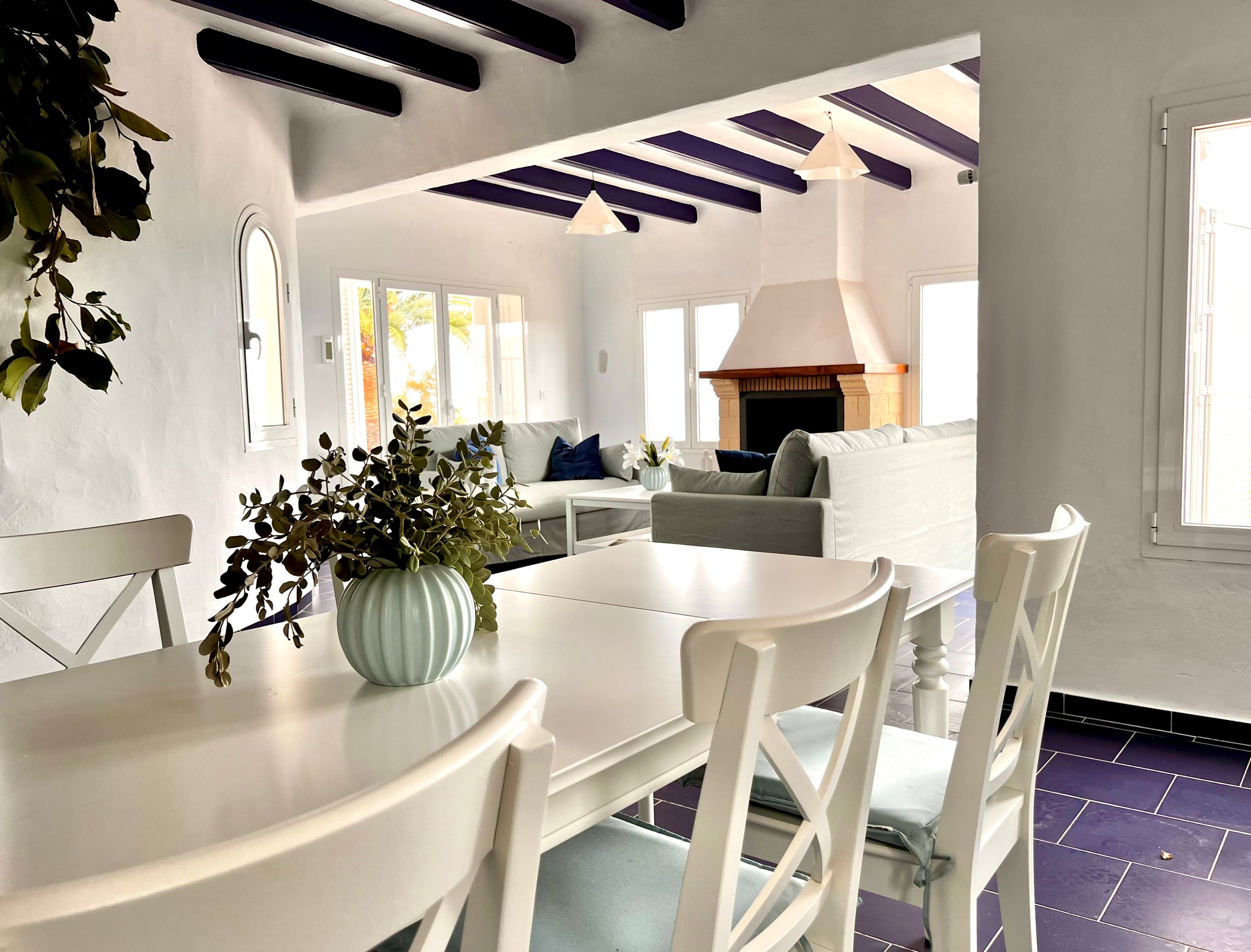 Bright modern dining area with white furniture, exposed beams, and a potted plant centerpiece in a coastal-style interior.