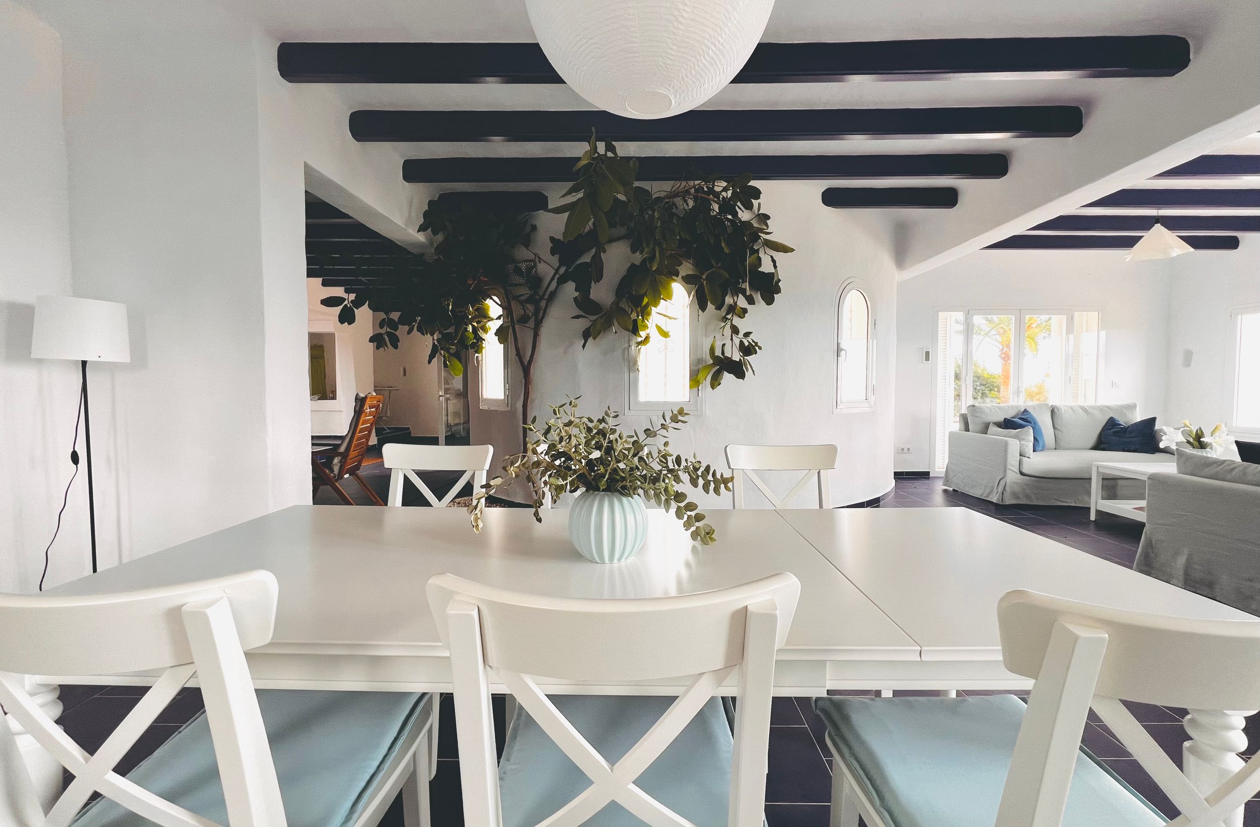 Modern white dining area with exposed ceiling beams, cross-back chairs, and indoor plants in a bright open space.