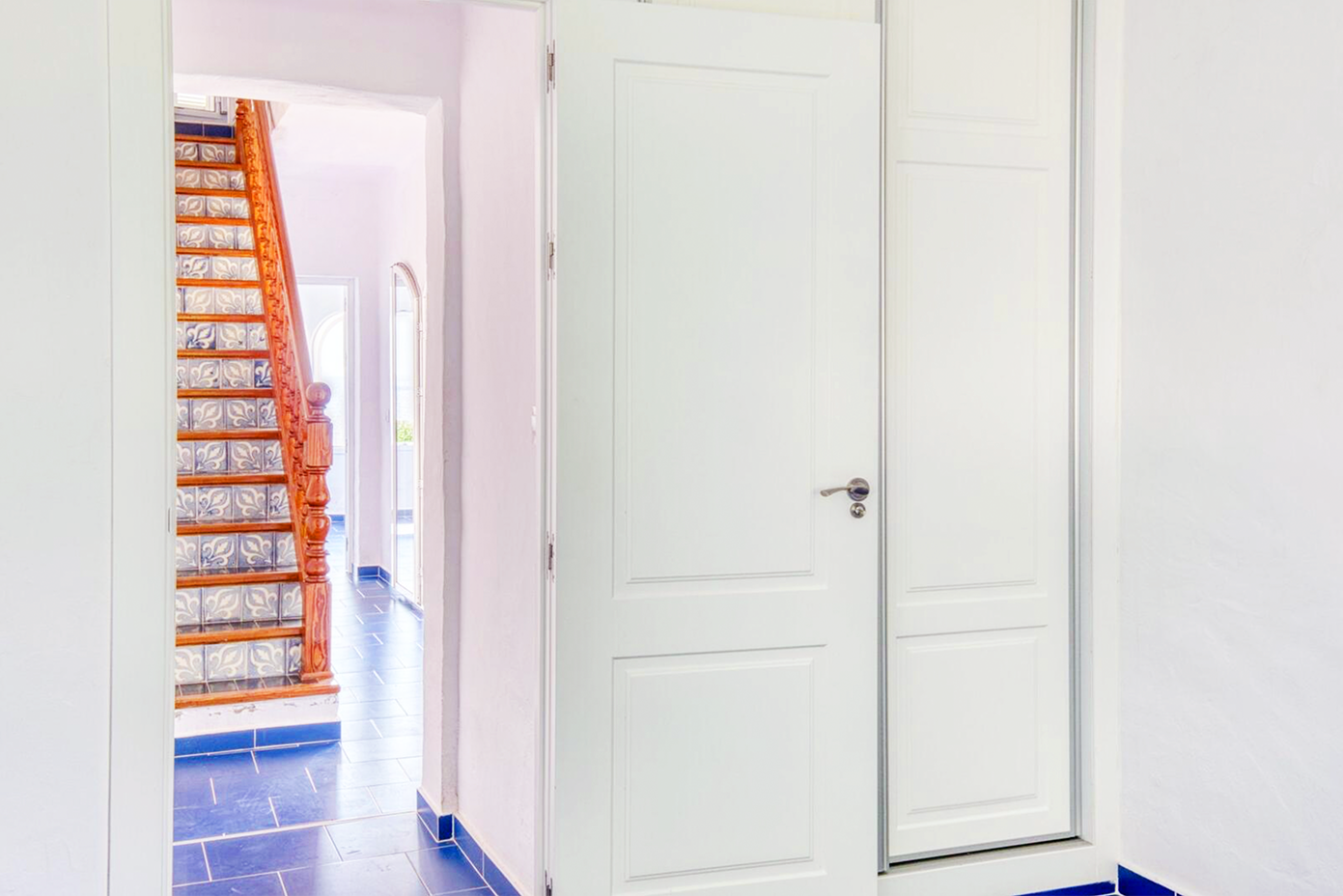 White hallway with orange wooden stairs and blue floor tiles leading to closed doors.