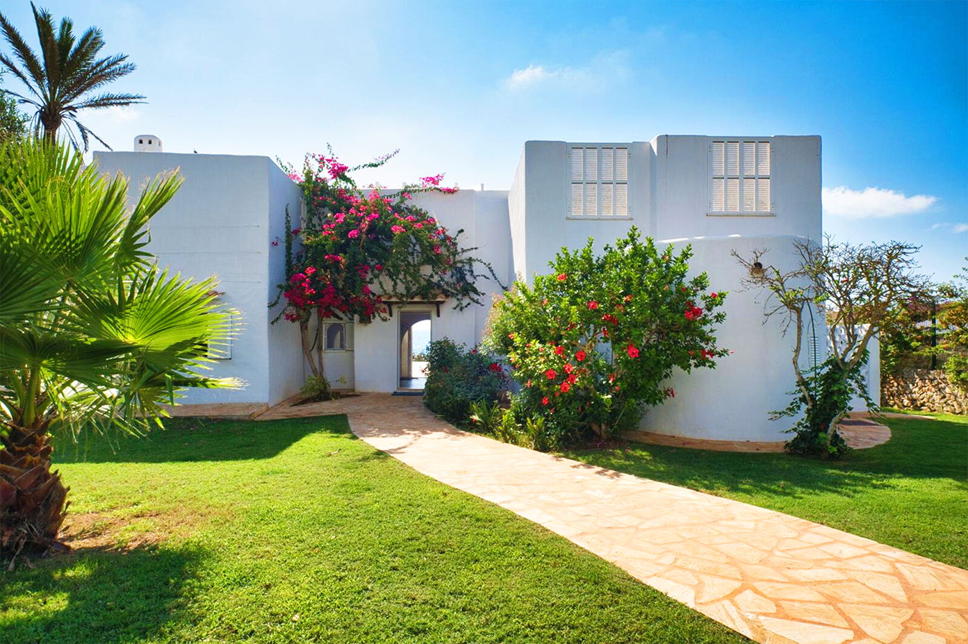 Modern white Mediterranean villa with vibrant flowering plants and palm trees along a stone pathway.