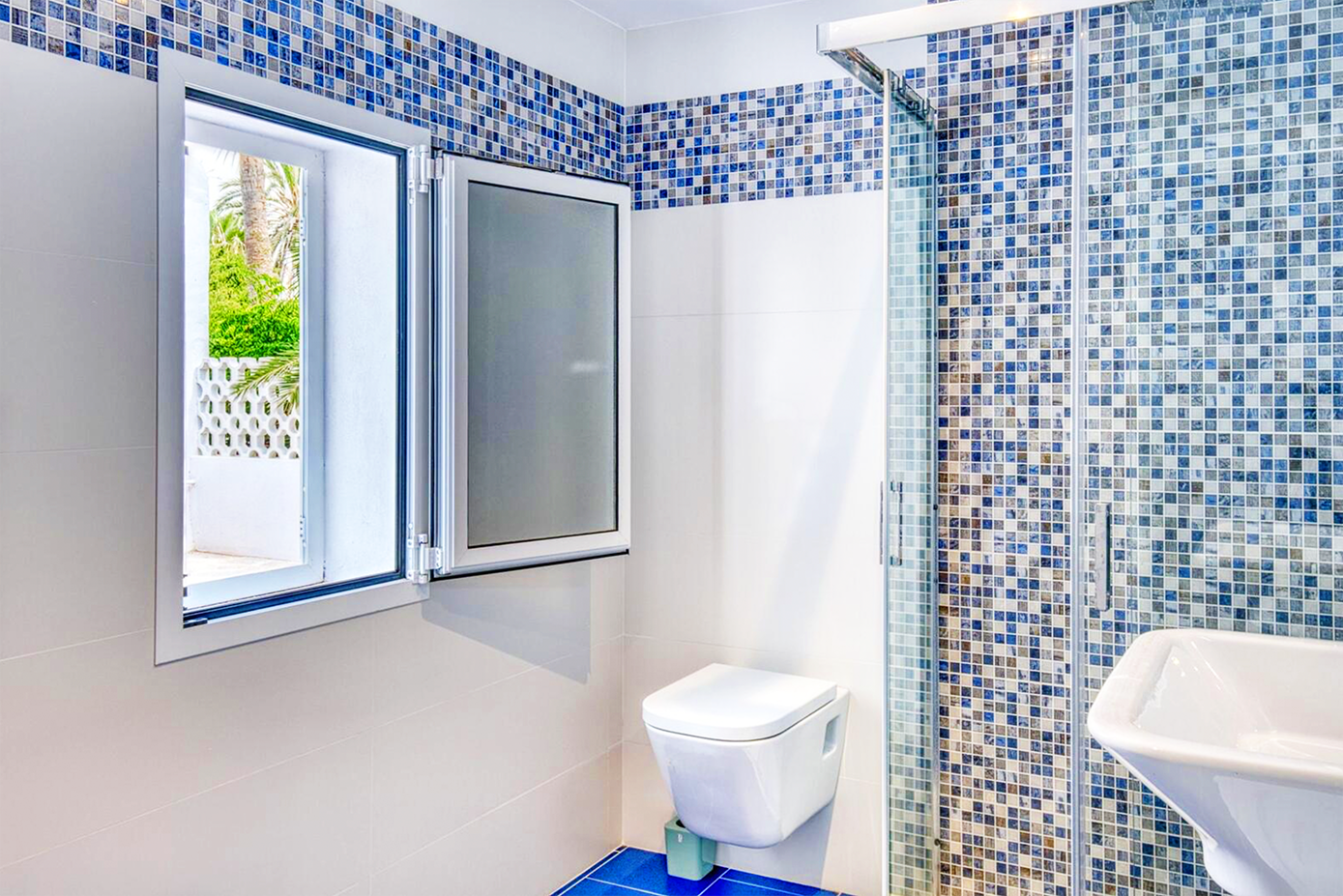 Modern bathroom with blue mosaic tile walls, white fixtures, and a window letting in natural light.