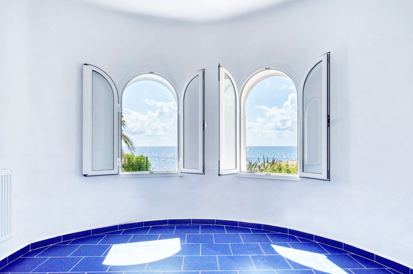 White room with arched windows overlooking the Mediterranean Sea and blue tile flooring.