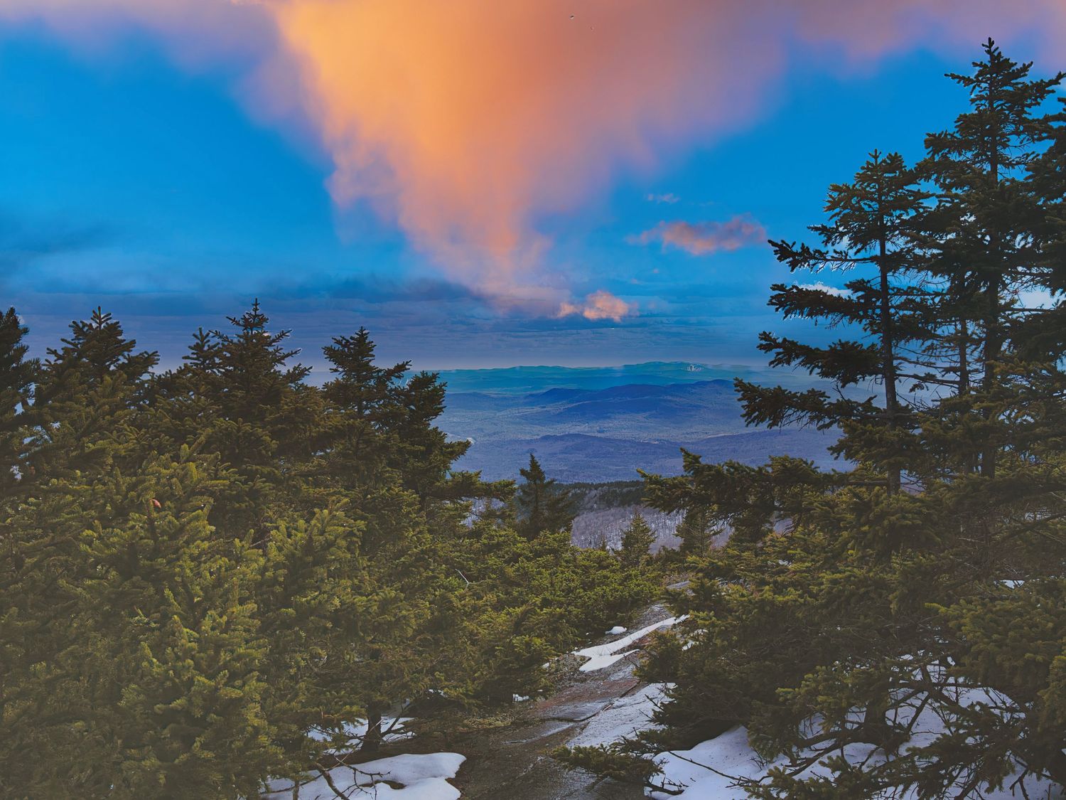 Stunning mountain sunset view through snow-dusted evergreen trees with vibrant orange and blue sky.