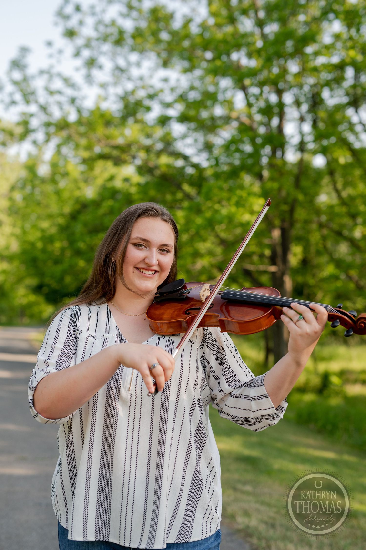 Musician plays violin outdoors on sunny path with green foliage in background.
