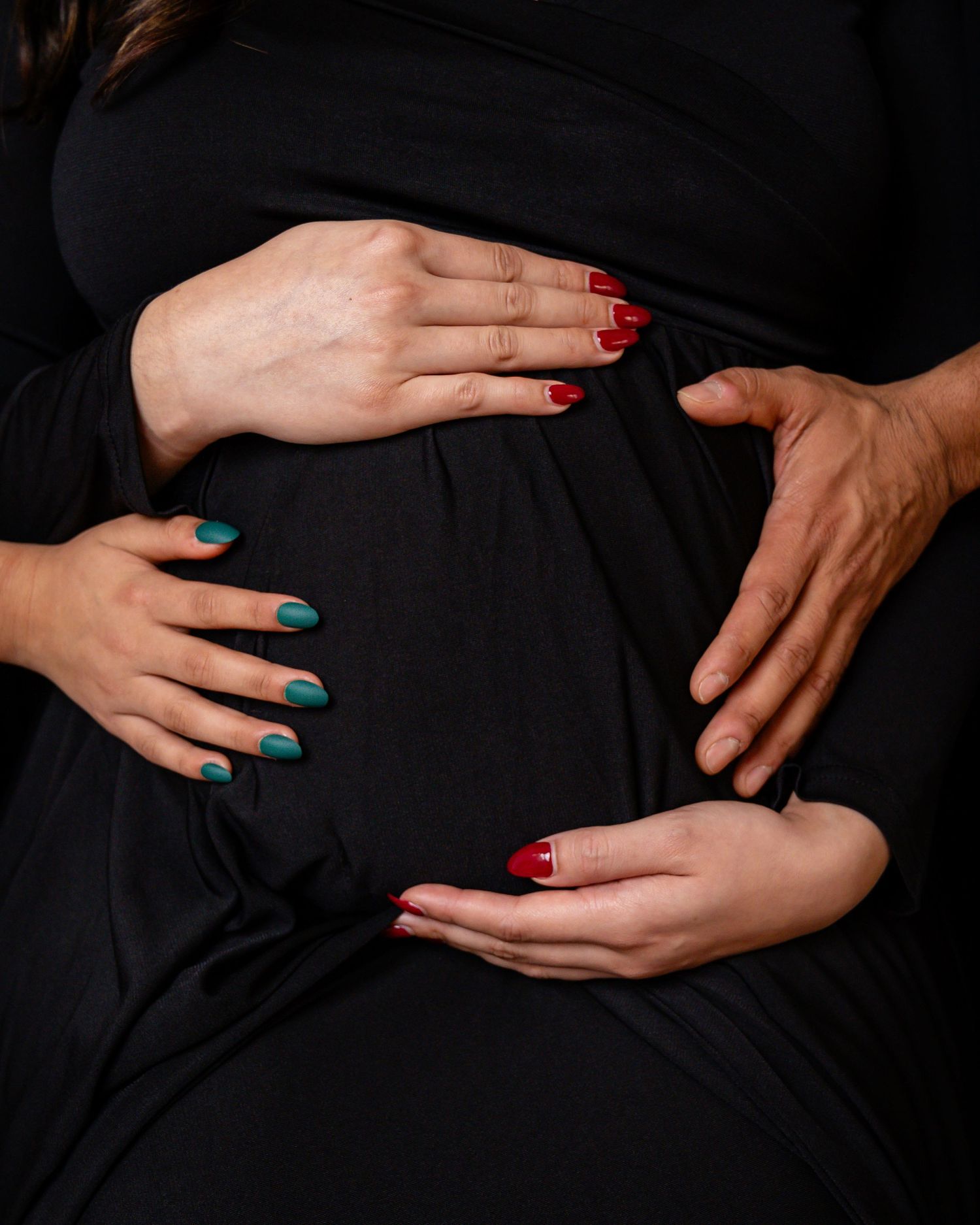 Close-up of hands with colorful nails cradling pregnant belly against black fabric.