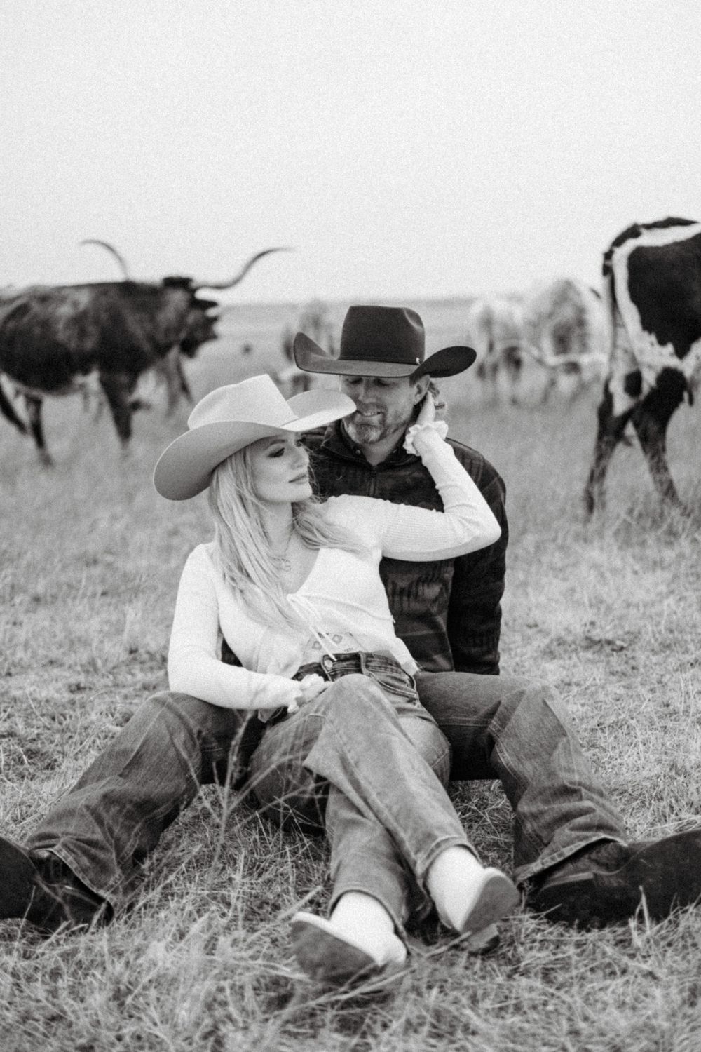 Black and white photo of western couple relaxing in field with longhorn cattle.
