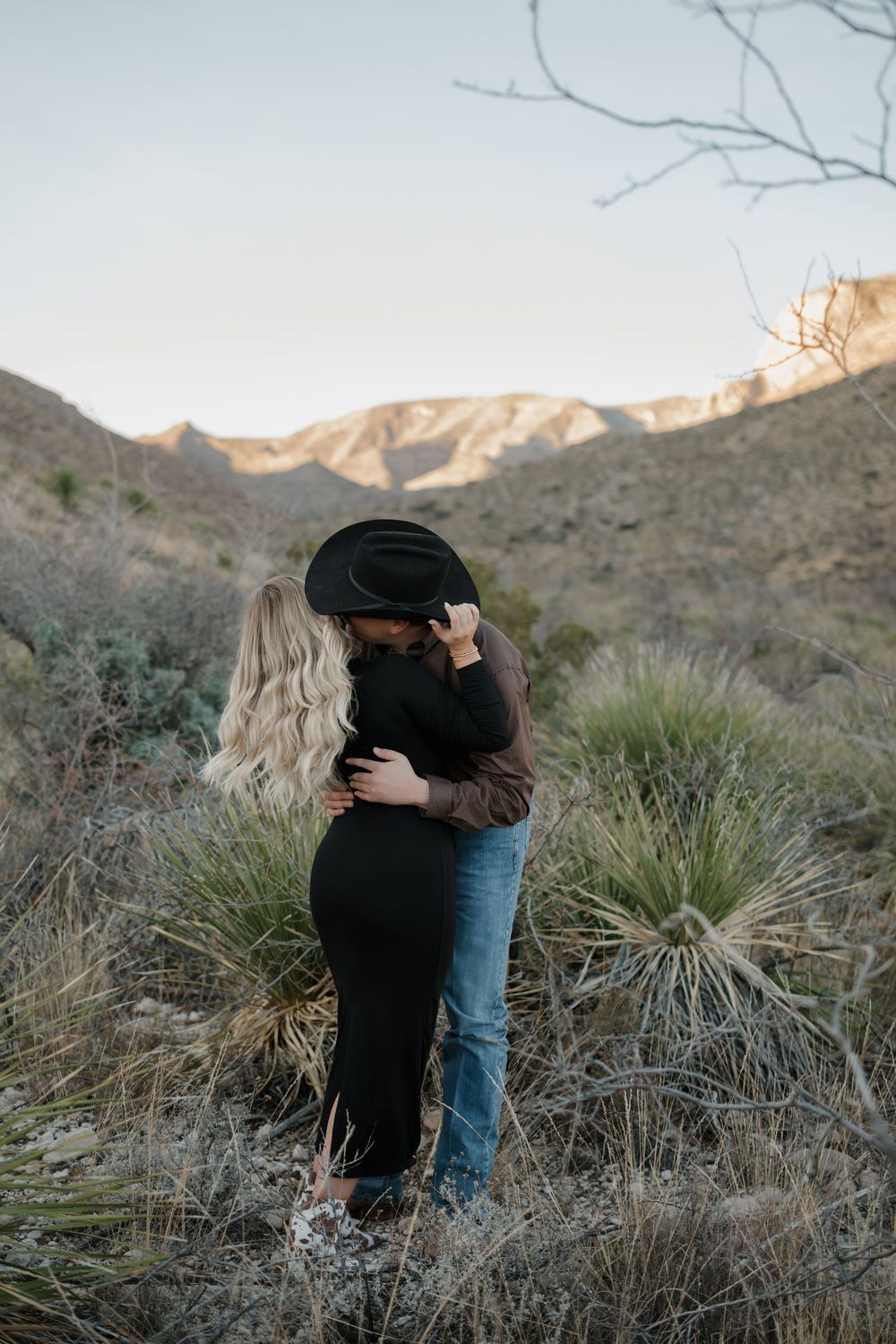 A couple embraces in a desert landscape with mountains and desert plants in the background during sunset.