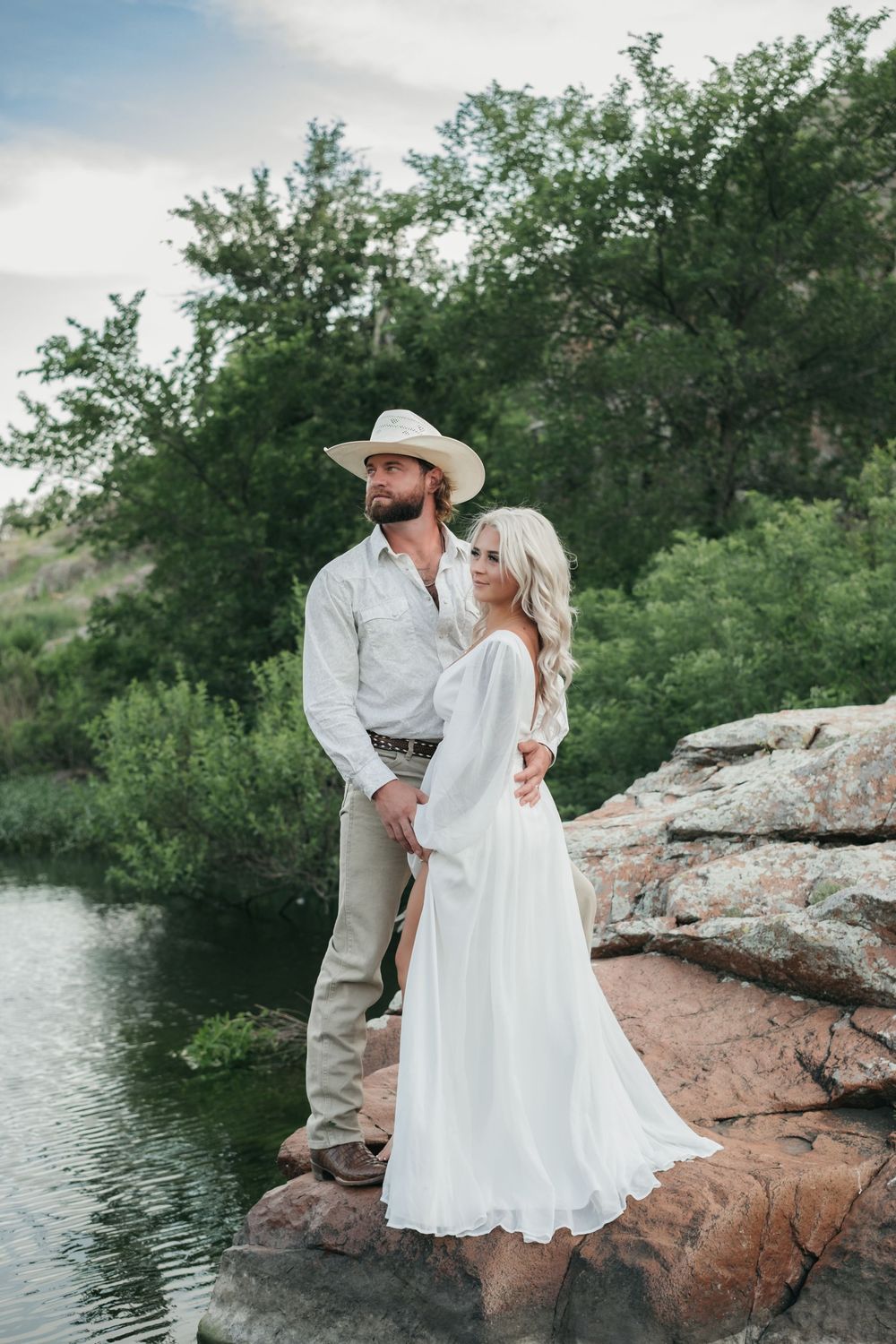 Couple in western attire standing on rocky outcrop overlooking tranquil lake.