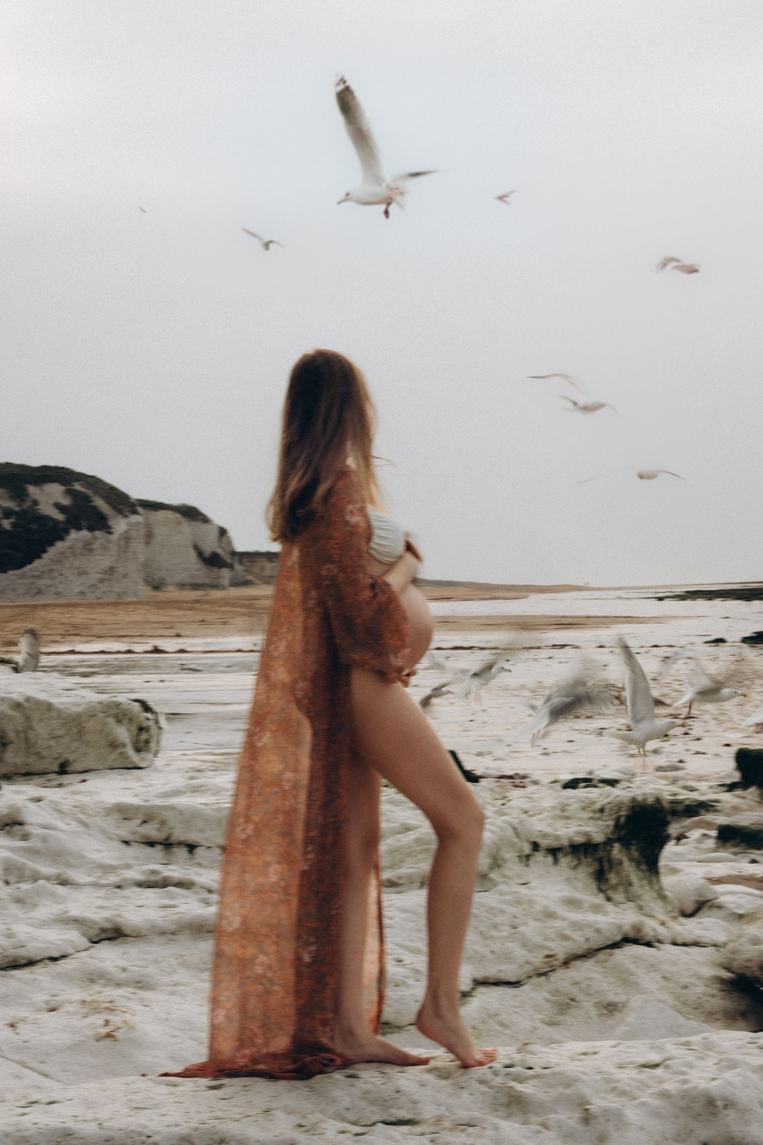 A pregnant woman standing on the beach while seagulls fly in the background