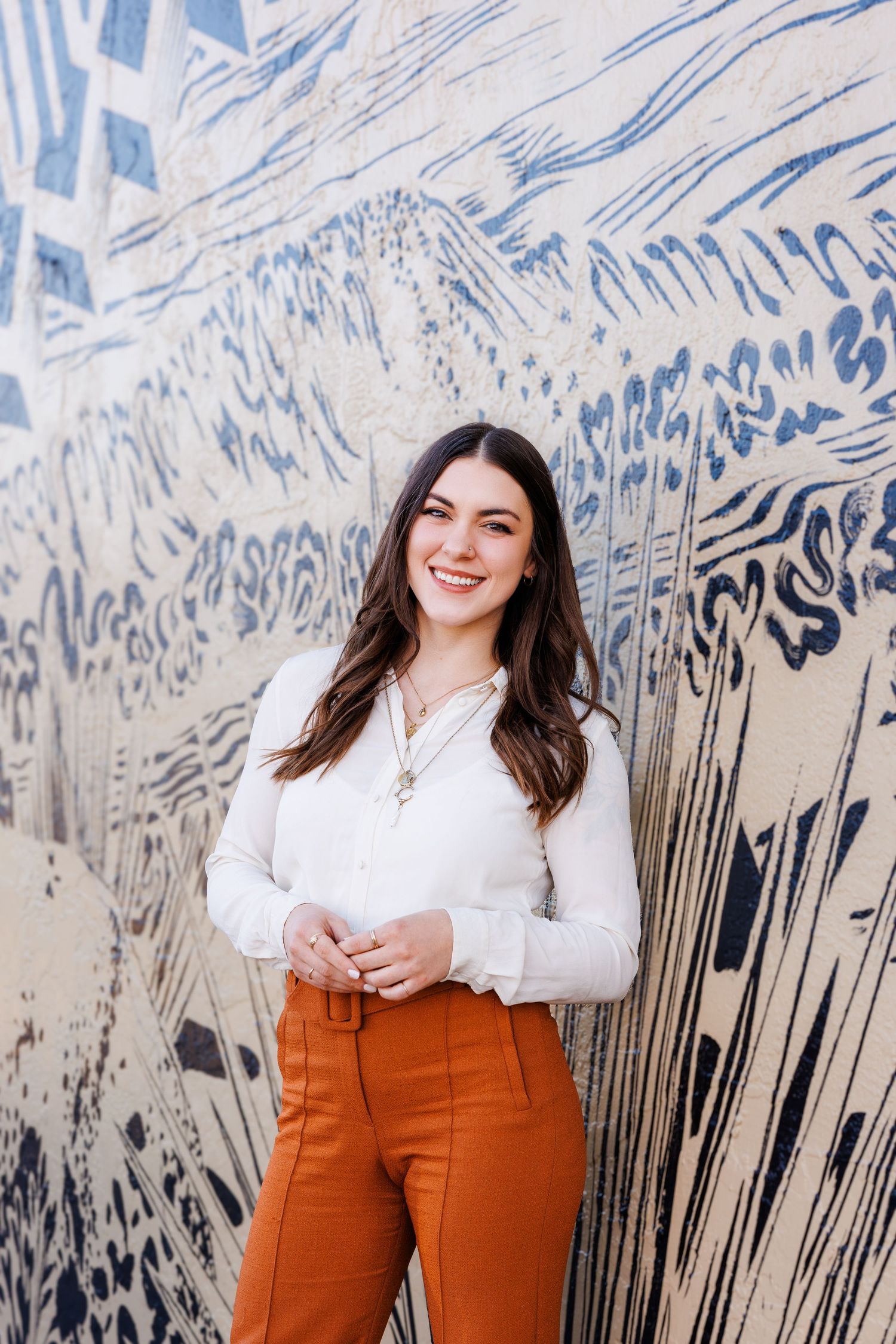 Woman in white top and orange pants poses against artistic blue wall mural.