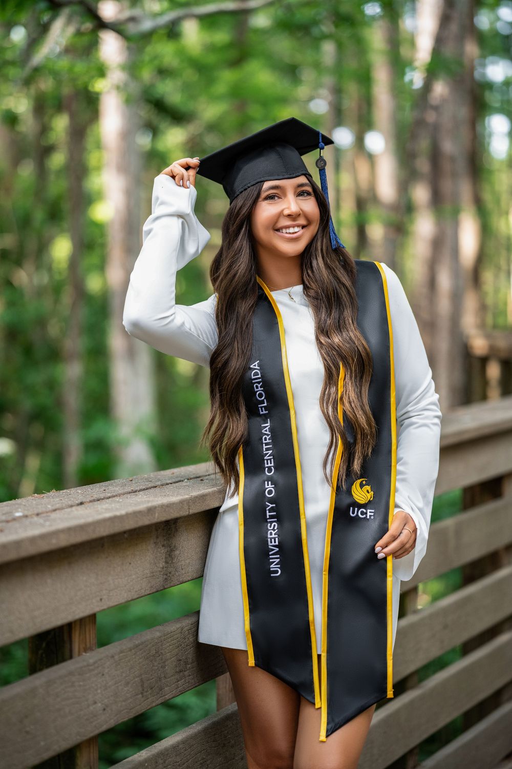 Graduate in black cap and gown with gold stole poses in woodland setting against wooden railing.