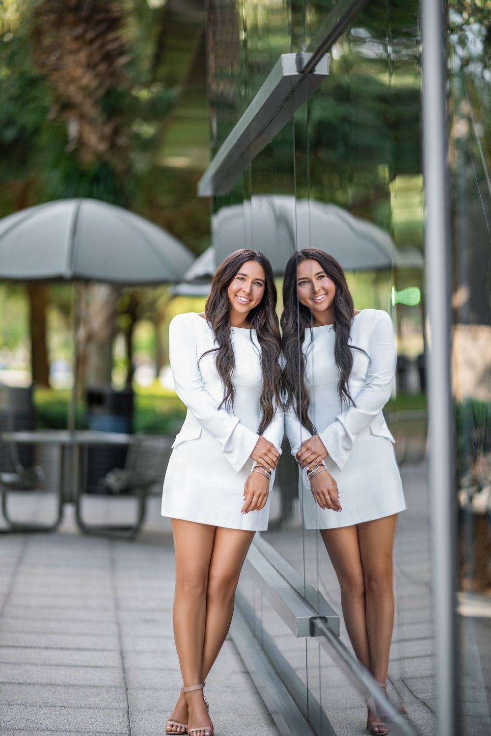 A woman in a white blazer poses playfully against a glass wall reflection in an urban setting.
