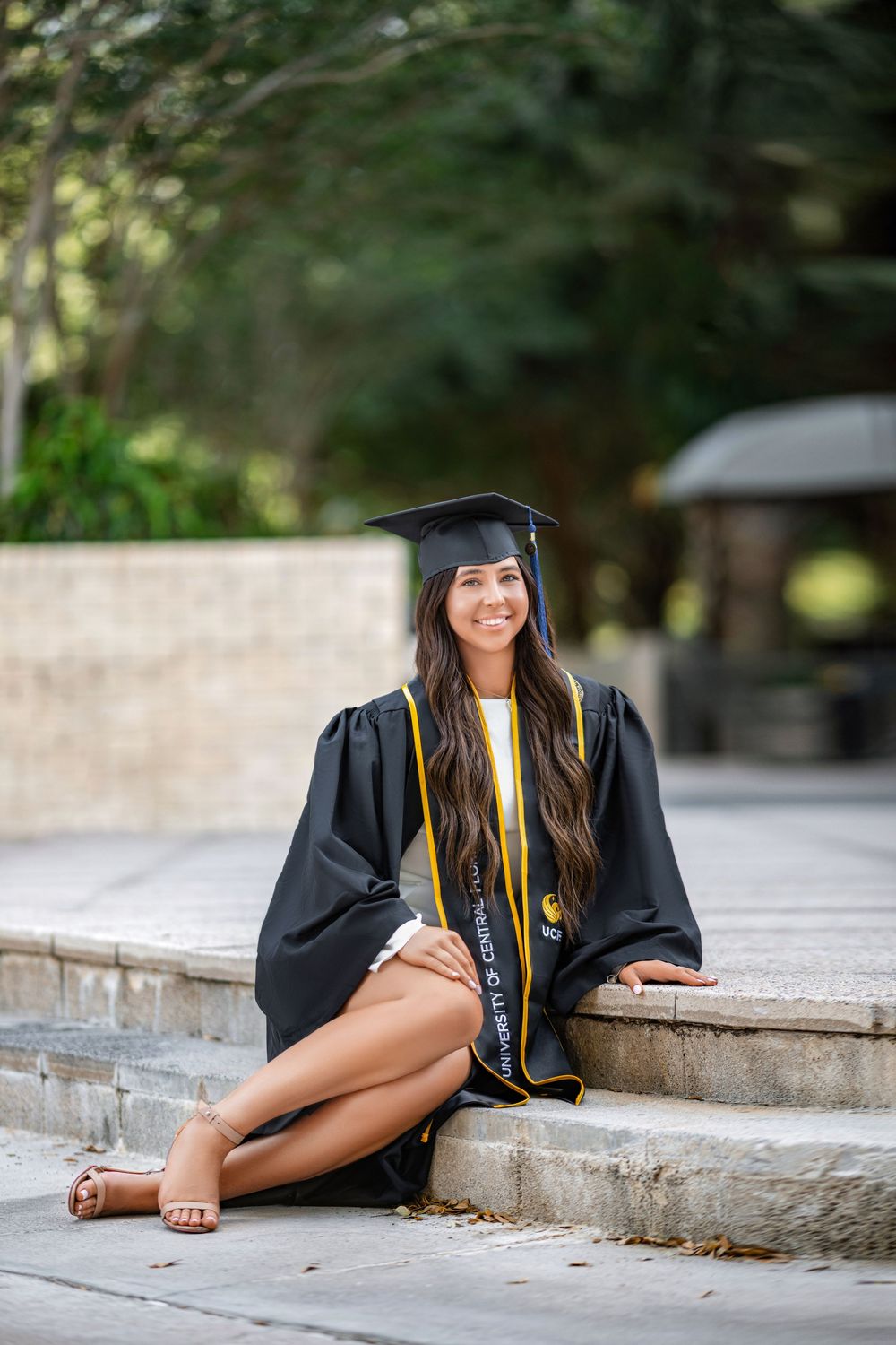 Graduate in cap and gown sitting on concrete steps outdoors during graduation photo session.