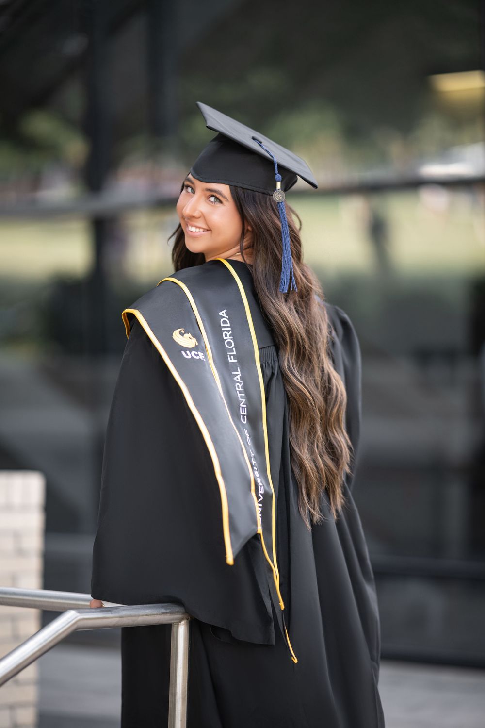 College graduate wearing black graduation cap and gown with gold trim poses outdoors during graduation photoshoot.
