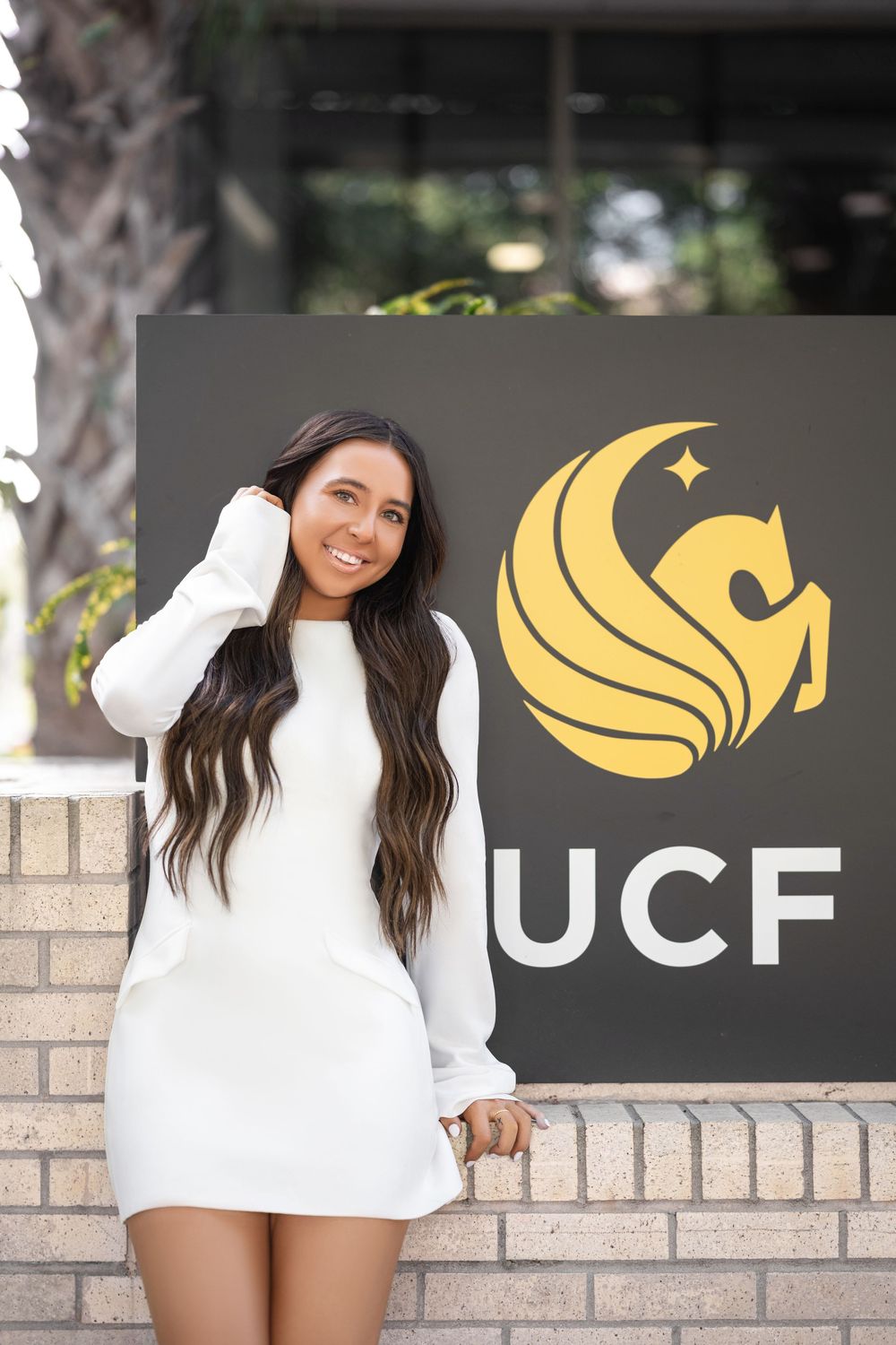 A woman in a white sweater dress poses against a brick wall with a black and gold UCF logo.