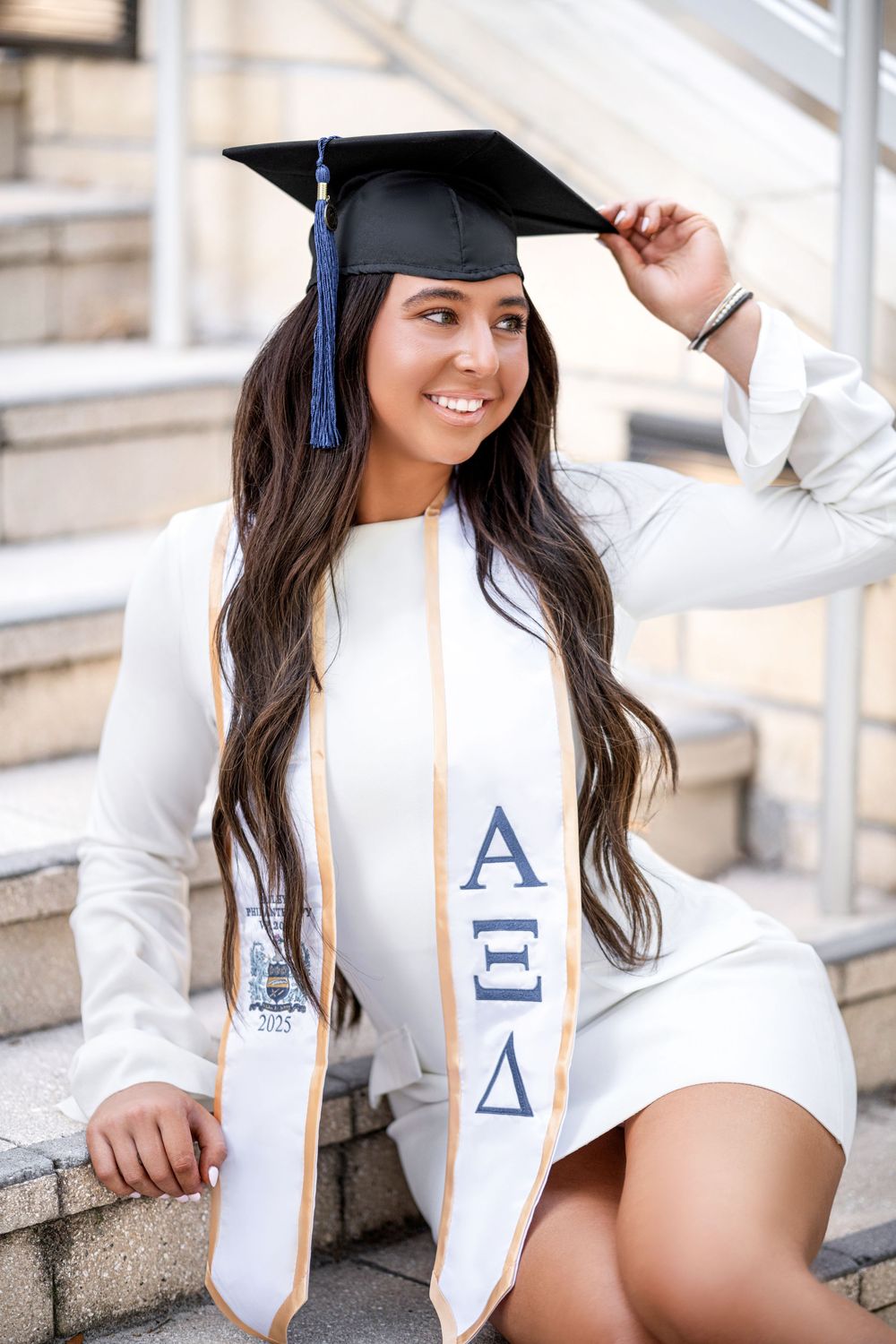 College graduate in white gown with graduation cap and stole poses on stone steps for portraits.