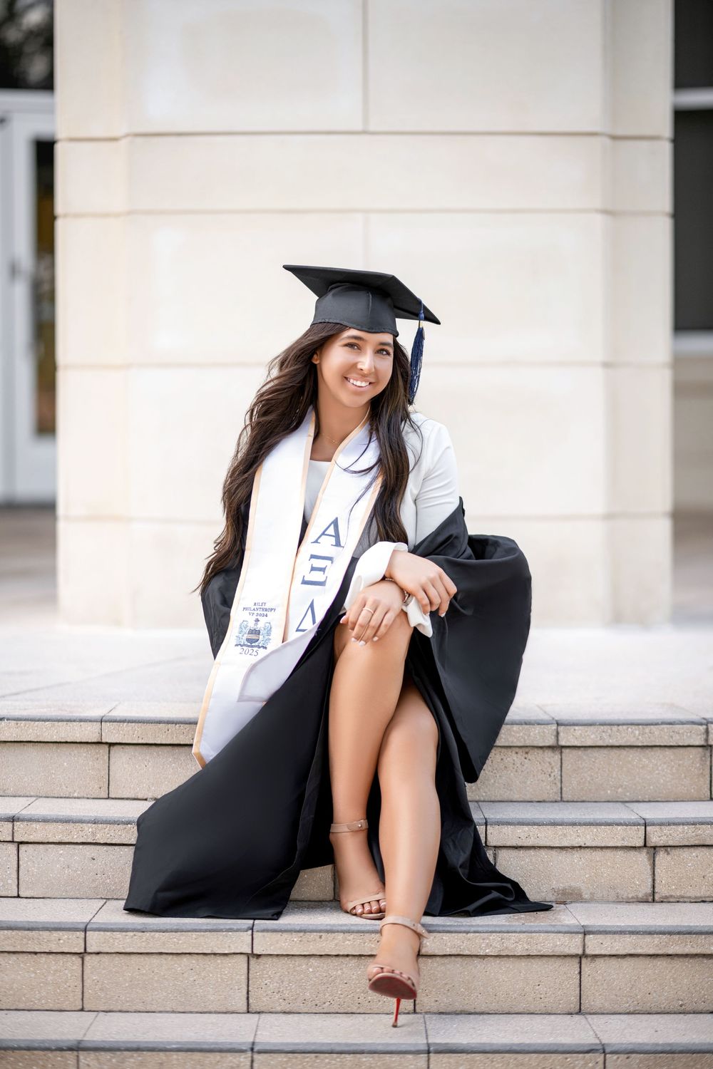 A graduate in cap and gown poses cheerfully on stone steps during an outdoor graduation photo session.