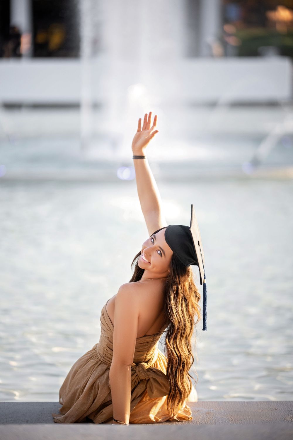 Graduate in cap and gown poses elegantly by water fountain during golden hour graduation photoshoot.
