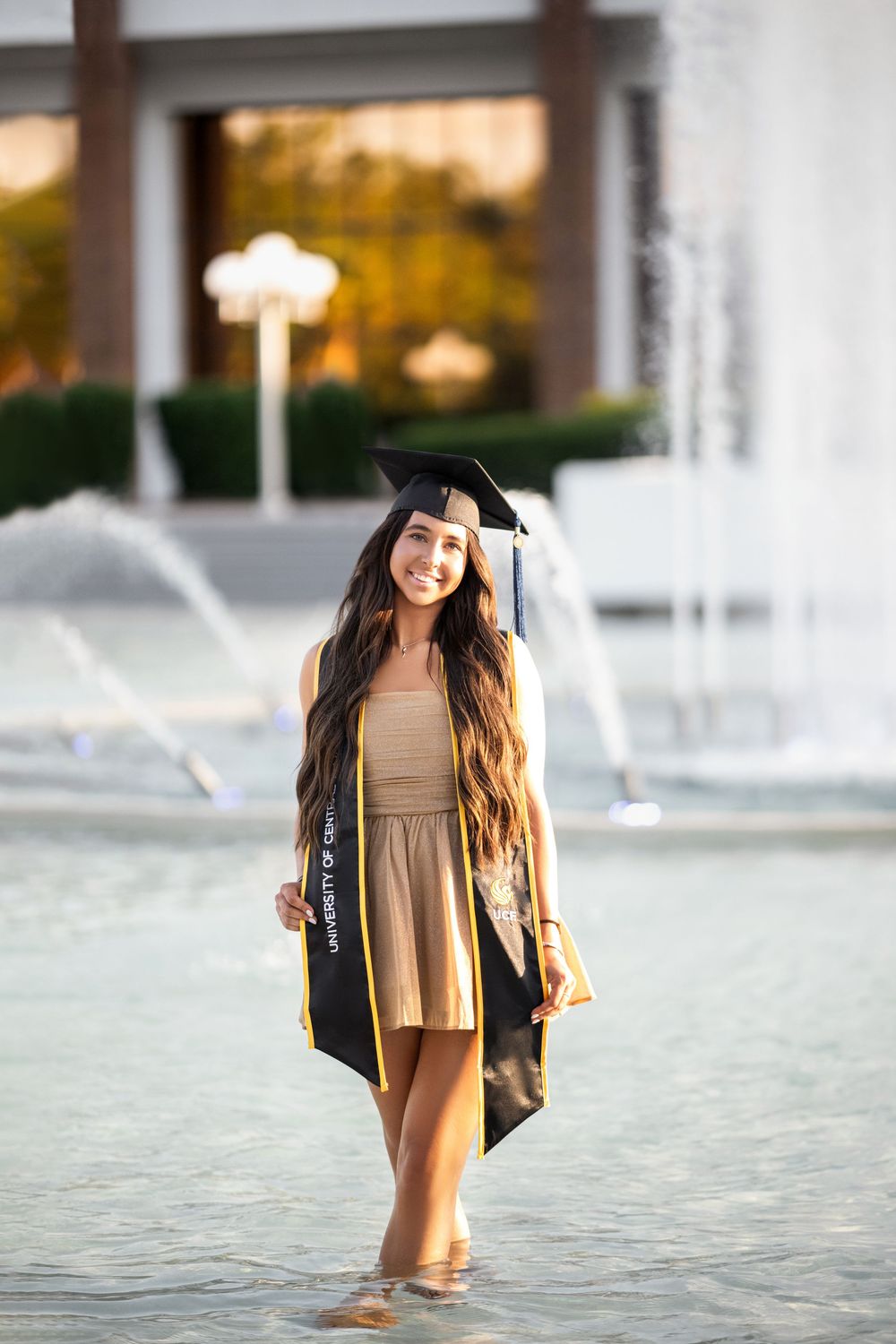 Graduate in a black cap and gown stands in a reflective fountain for graduation photos at sunset.