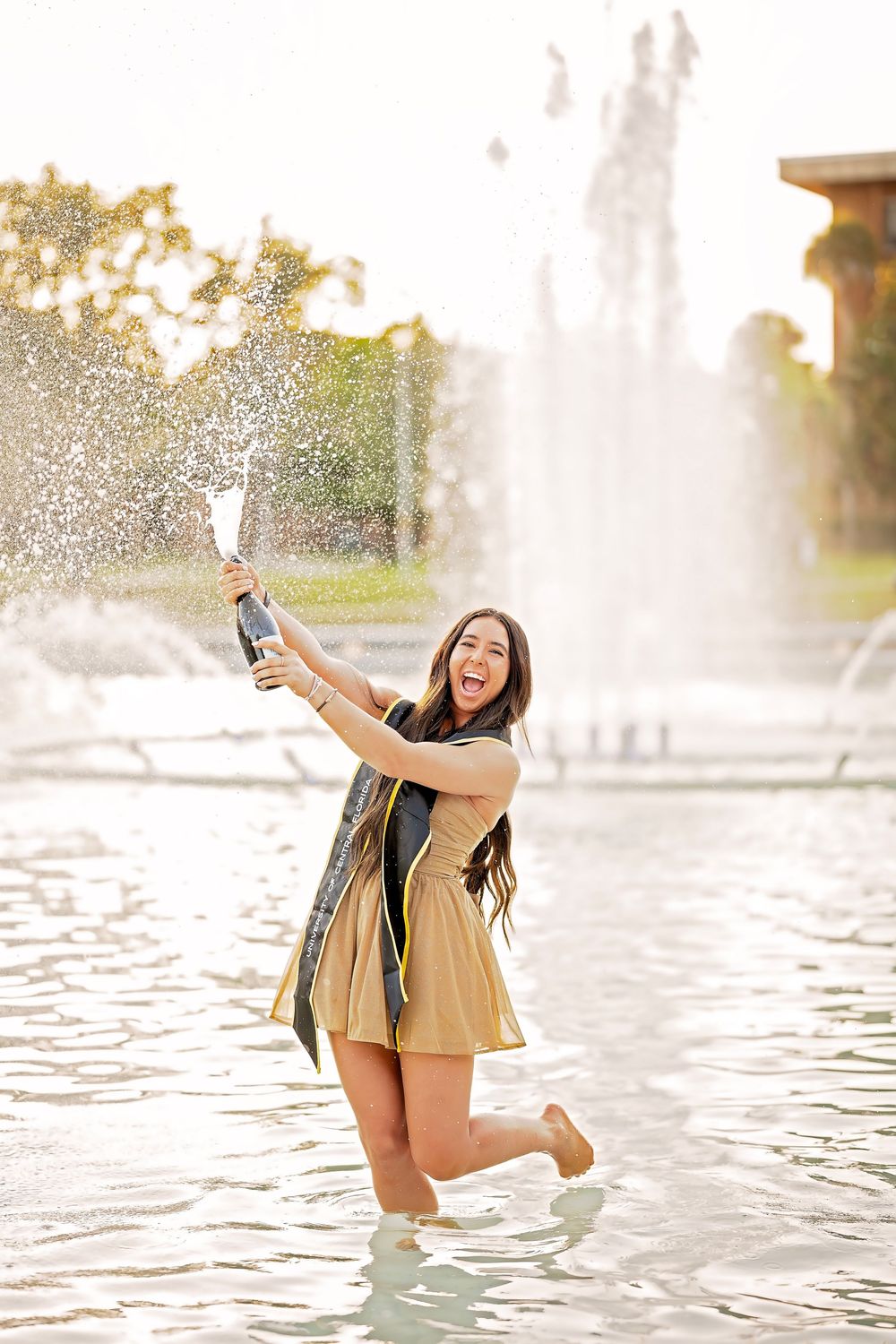 A joyful person in a tan dress splashes in a fountain while holding up a beverage on a sunny day.