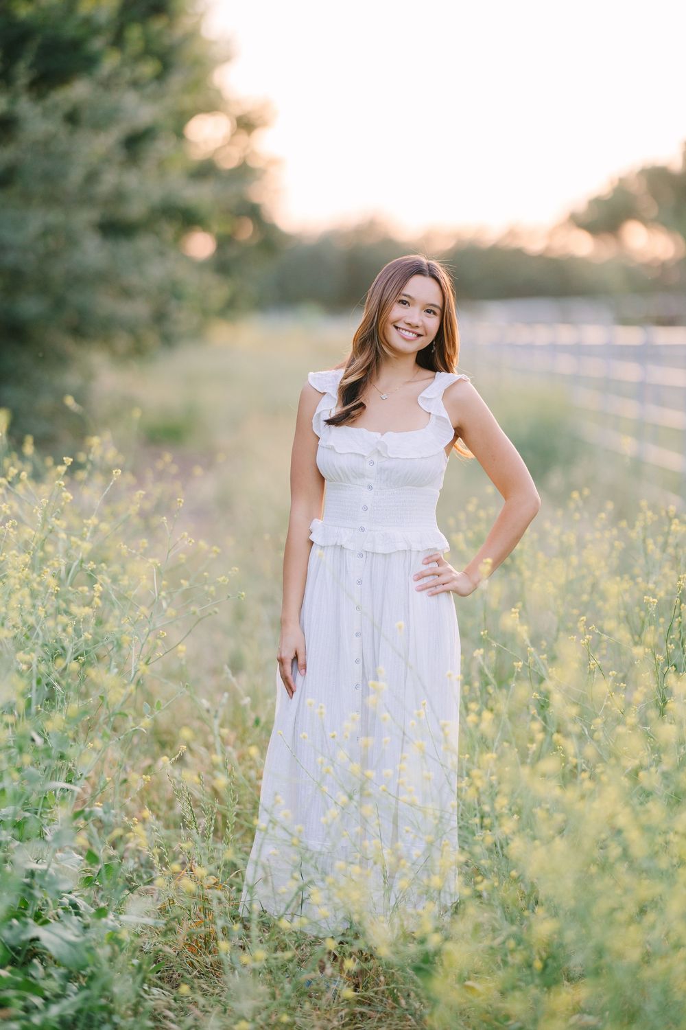 Young woman in white ruffle dress smiling in golden hour sunlit field with wildflowers and fence.