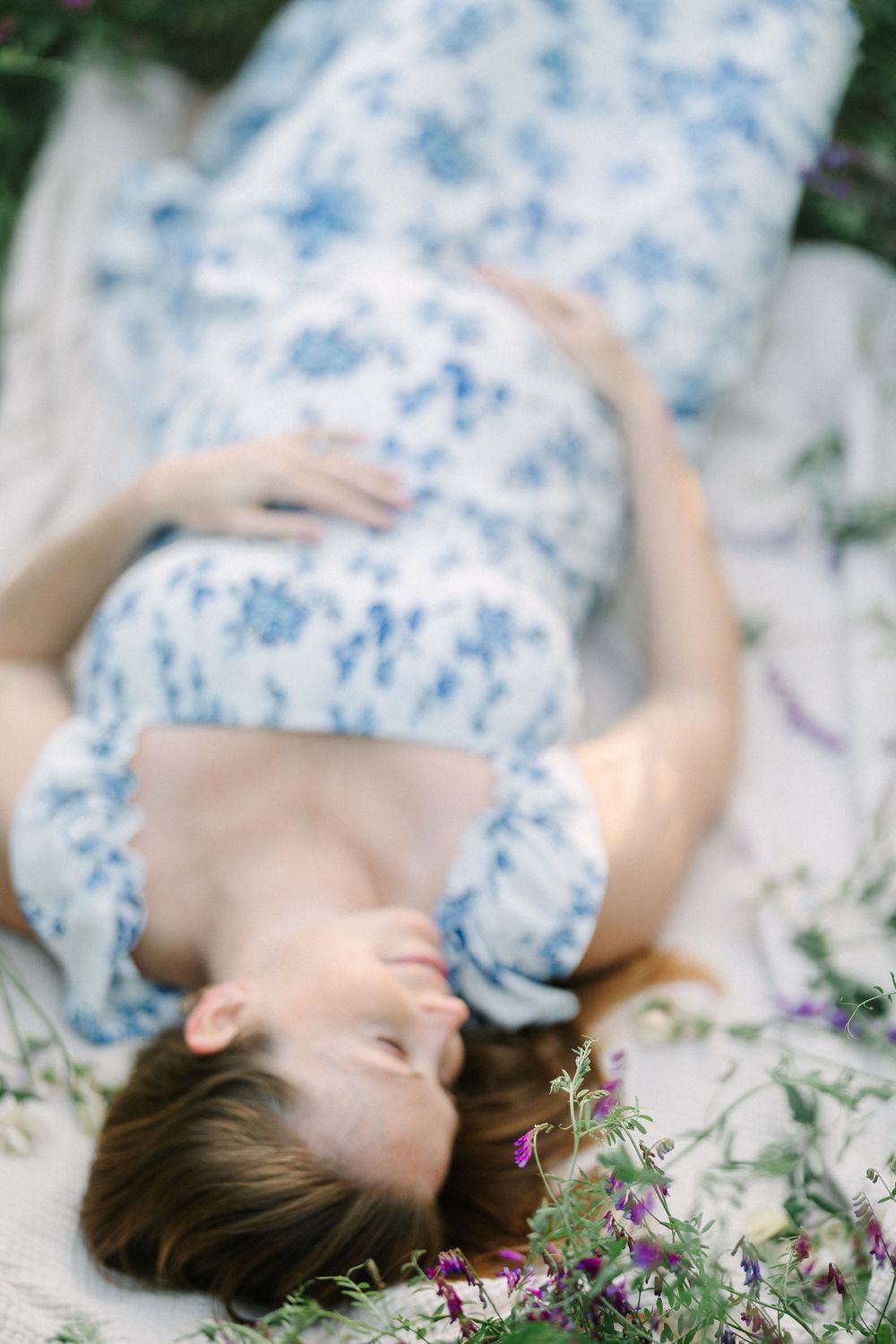 Pregnant woman in blue floral dress lying on blanket outdoors, cradling baby bump surrounded by wildflowers.