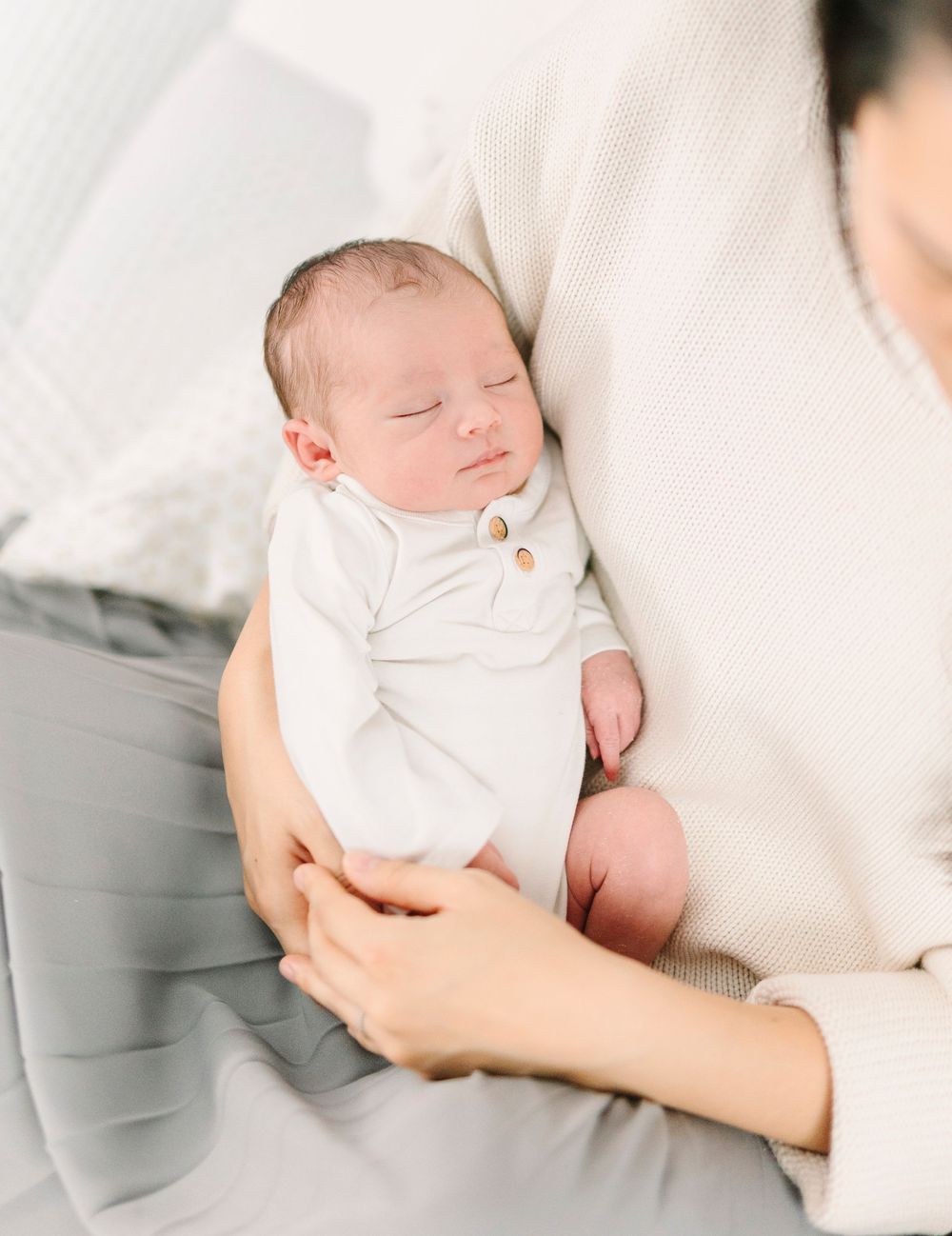 A newborn baby in white clothing rests peacefully on light gray fabric.