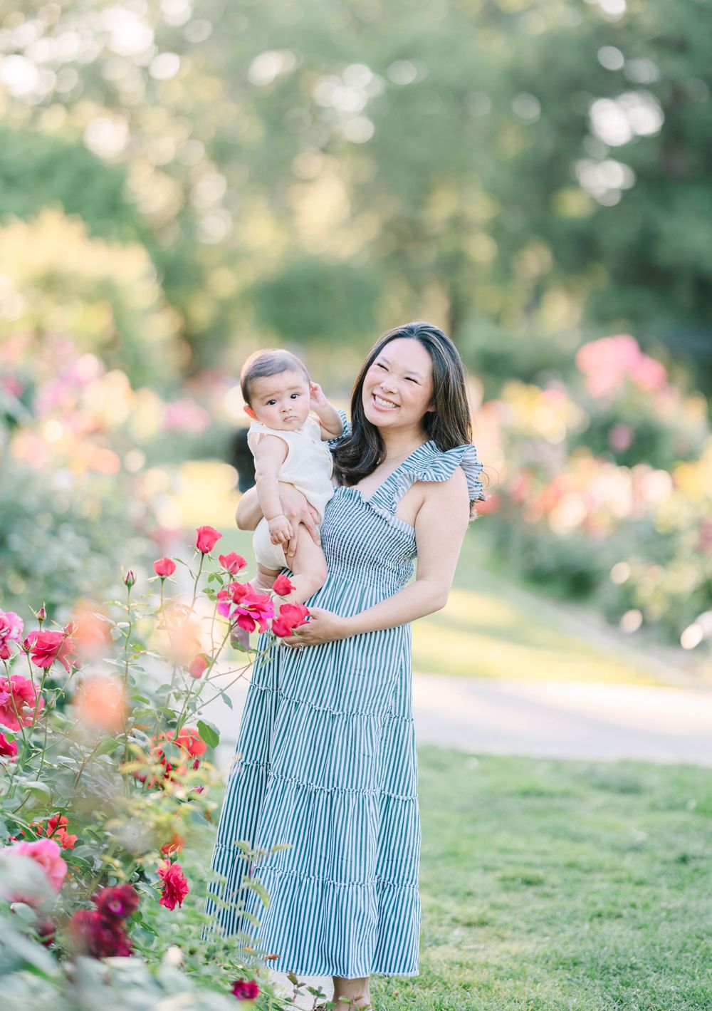 A smiling woman in a blue striped dress holds her baby among blooming pink roses in a lush garden.