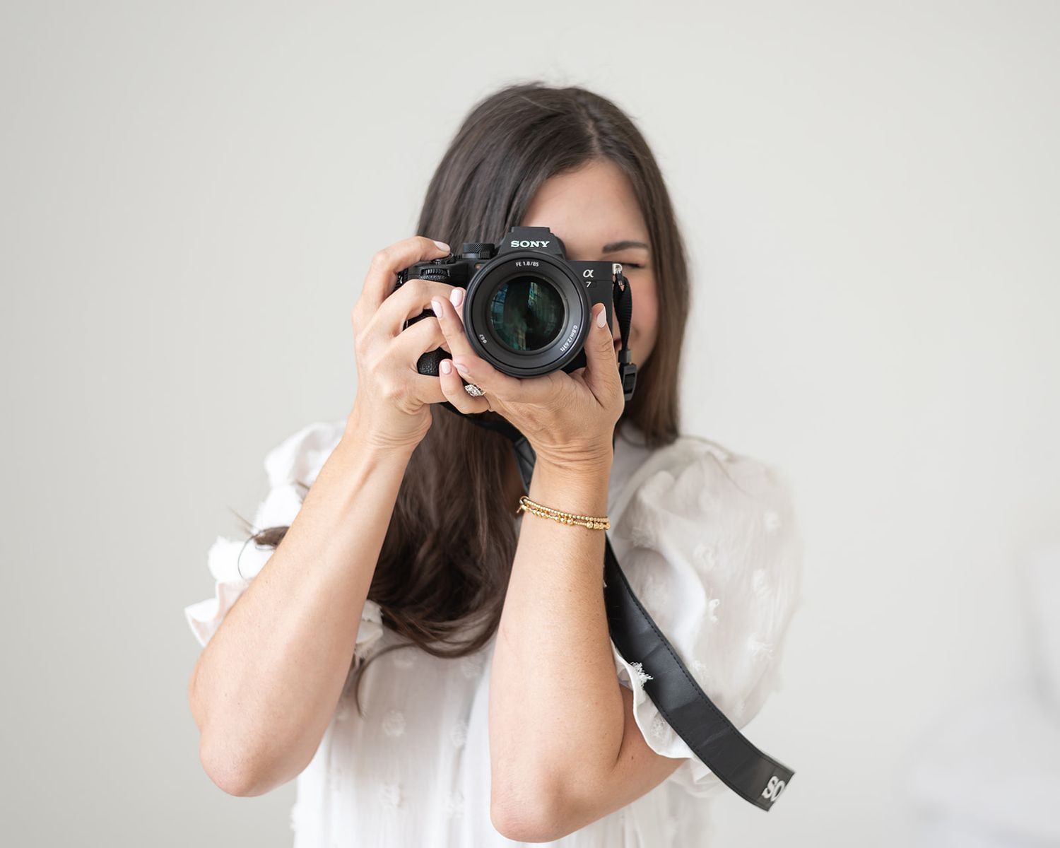 A photographer in a white blouse holds up a professional DSLR camera with a large lens against a light background.